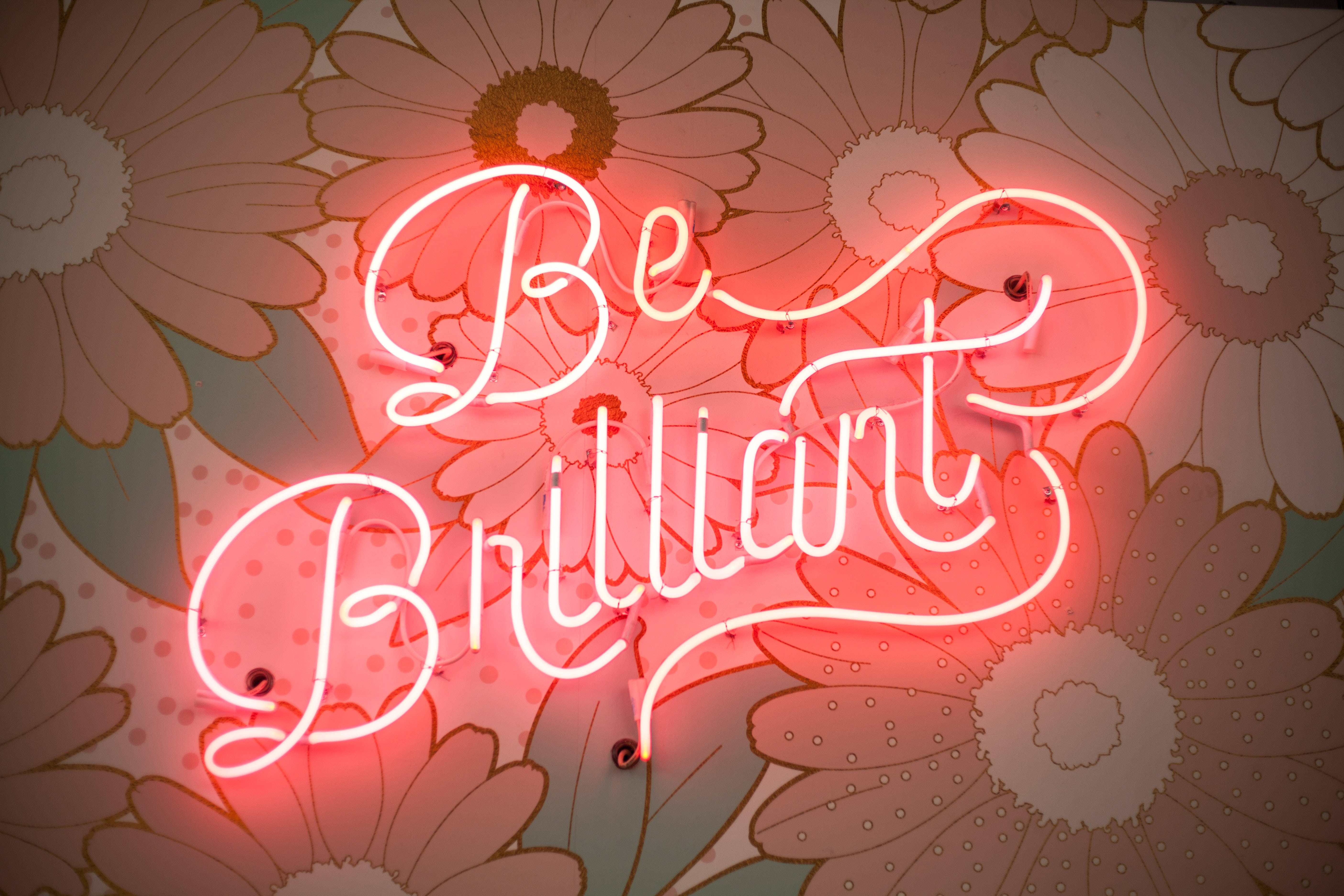 Cool 4K 桌布 壁紙 — 一個發光的紅色霓虹燈牌，上面寫著「Be Brilliant」，在復古花卉壁紙背景下閃耀。