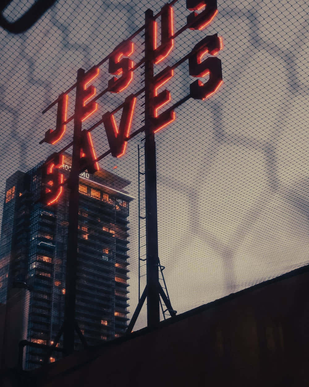 Jesus Saves 桌布 壁紙 — 一個發光的紅色霓虹燈牌，上面寫著「JESUS SAVES」，在暮色城市天際線前高高聳立，前景有鐵絲網圍欄。