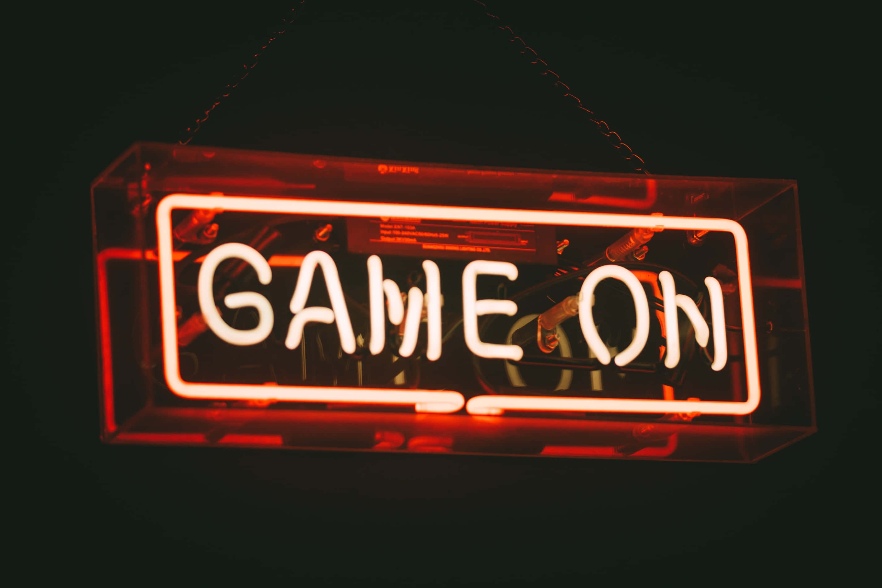 筆電 桌布 壁紙 — 一個發光的紅色霓虹燈牌，上面寫著 GAME ON，懸掛在黑暗中，將溫暖的光線投射到其透明的盒子框架上。