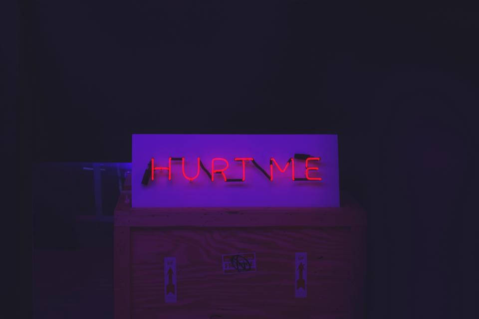 dark neon iPhone 桌布 壁紙 — 一個發光的紅色霓虹燈牌，上面寫著「HURT ME」，在黑暗的房間裡將紫色光暈投射在木箱上。