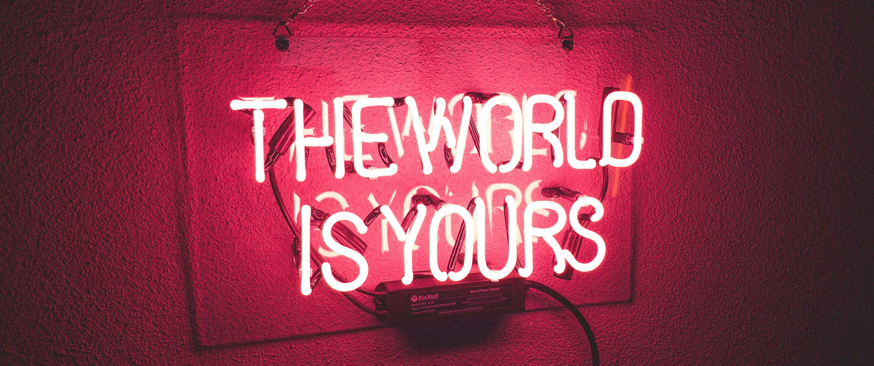 3440x1440 Neon 桌布 壁紙 — 一個發光的紅色霓虹燈牌，上面寫著「THE WORLD IS YOURS」，背景是紋理牆壁，投射出溫暖的環境光。