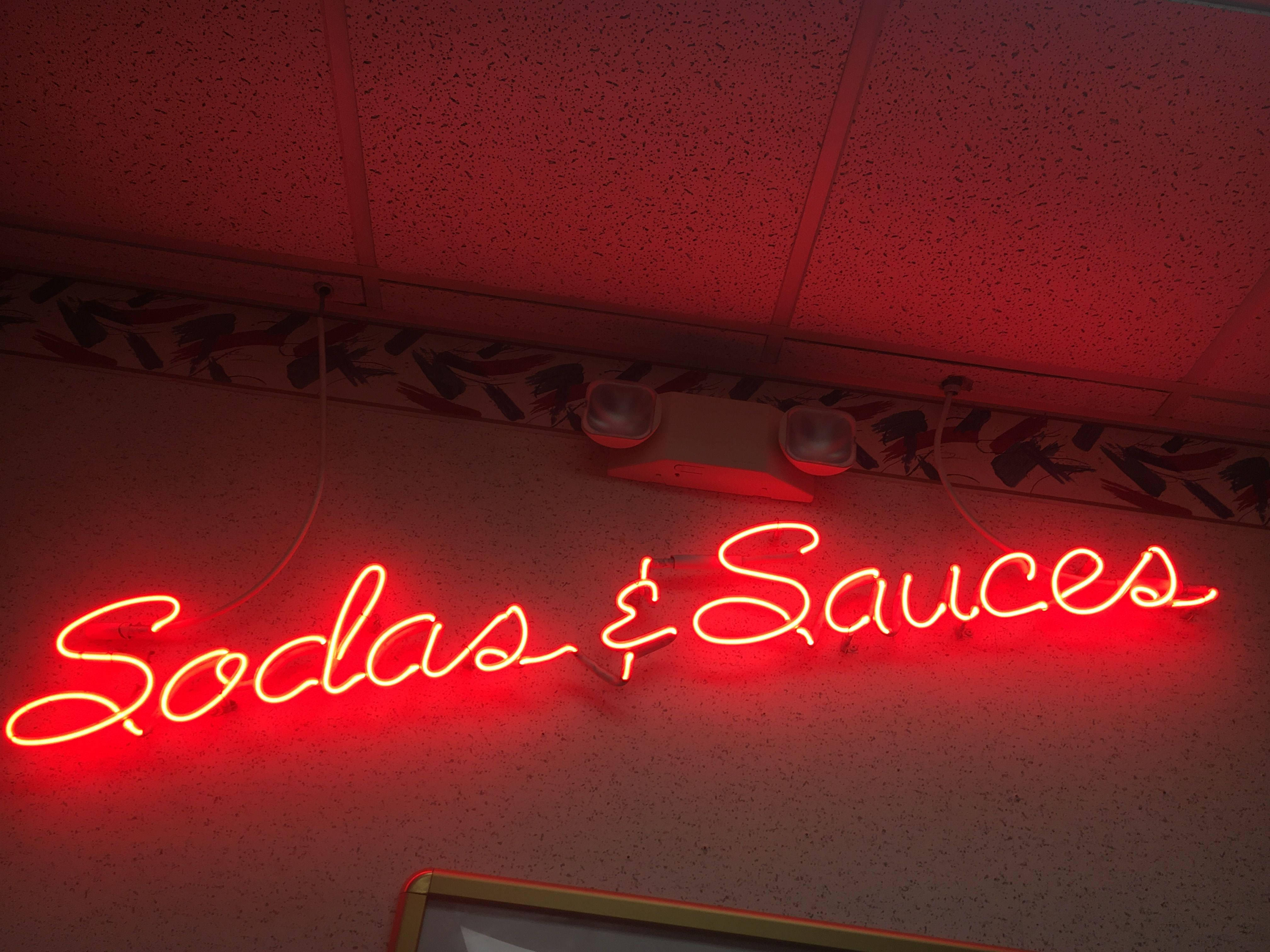 紅色動圖 桌布 壁紙 — 一個發光的紅色霓虹燈牌，上面寫著 Sodas & Sauces，在圖案邊框和螢光燈下的斑點牆上投射出溫暖的光線。