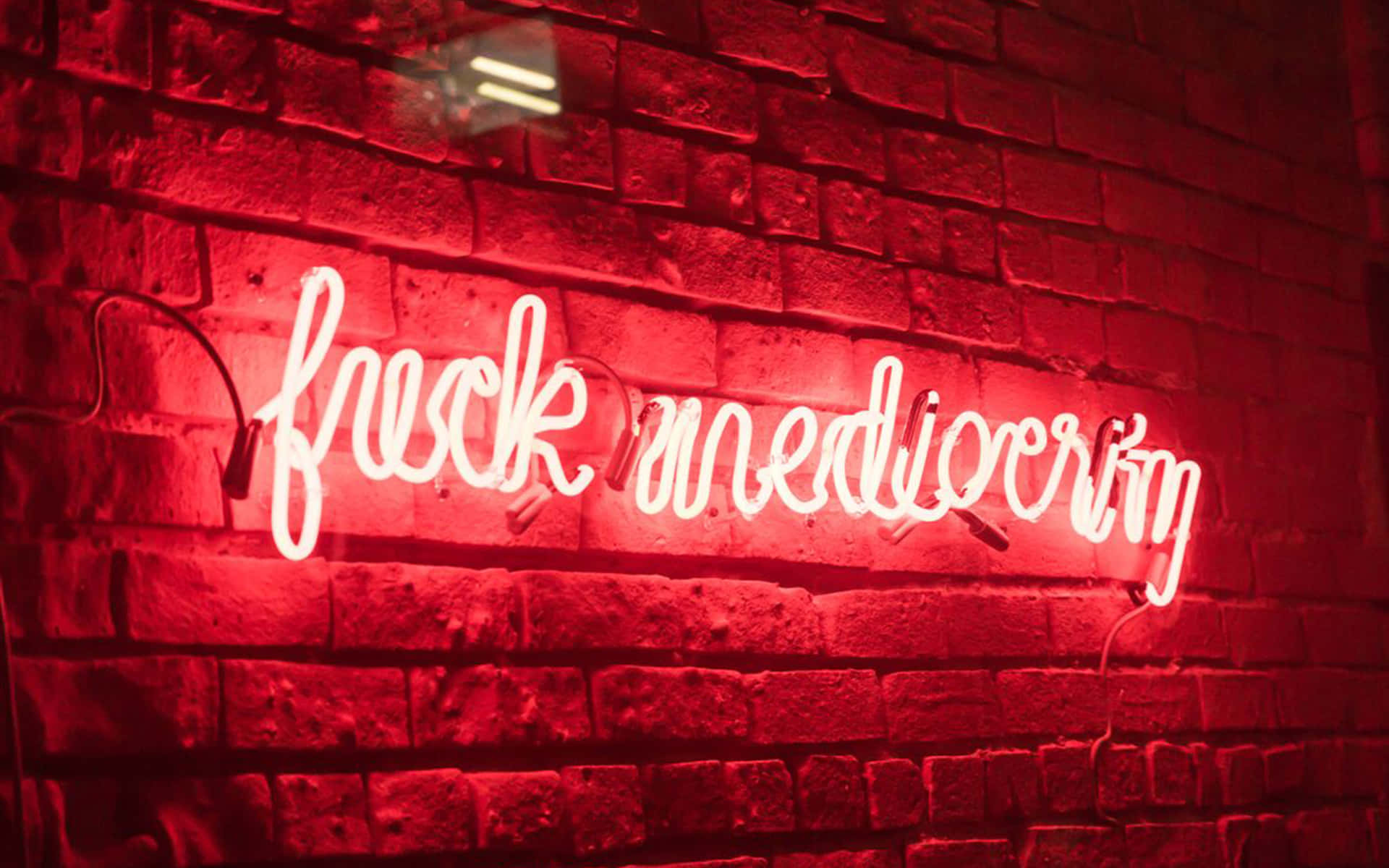 Neon Red Aesthetic 桌布 壁紙 — 一個發光的紅色霓虹燈牌，上面寫著「fuck mediocrity」，在昏暗的城市環境中，將大膽的光線投射在有紋理的磚牆上。
