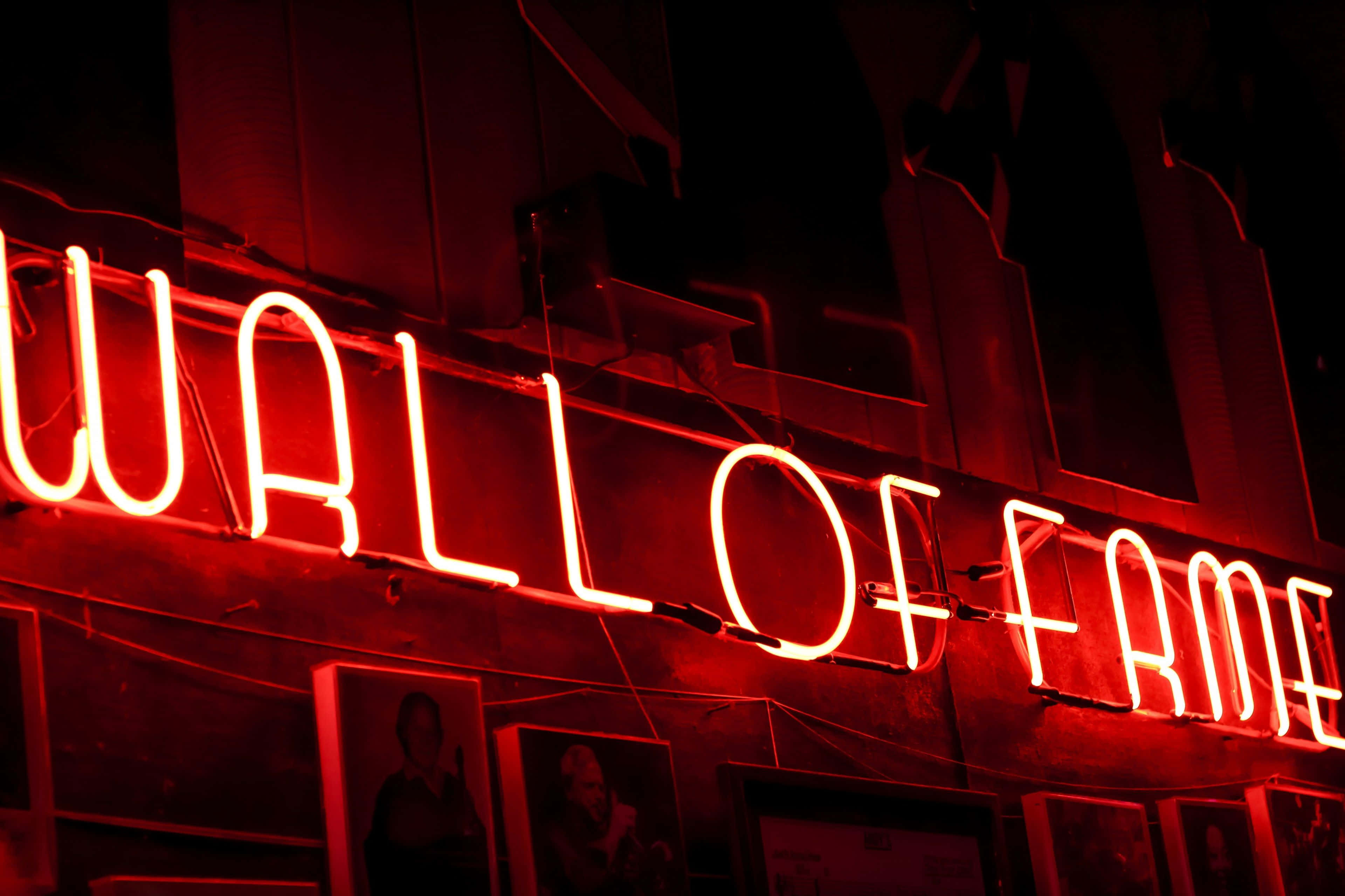 霓虹紅 桌布 壁紙 — 一個發光的紅色霓虹燈牌，上面寫著「WALL OF FAME」，照亮了下方掛有裱框照片的黑暗城市牆壁。