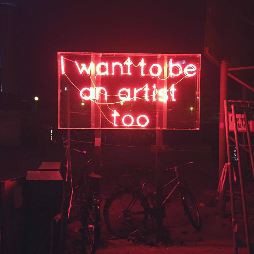 美學 桌布 壁紙 — 一個發光的紅色霓虹燈牌，上面寫著「I want to be an artist too」，照亮了夜晚黑暗的城市小巷，附近停著自行車。