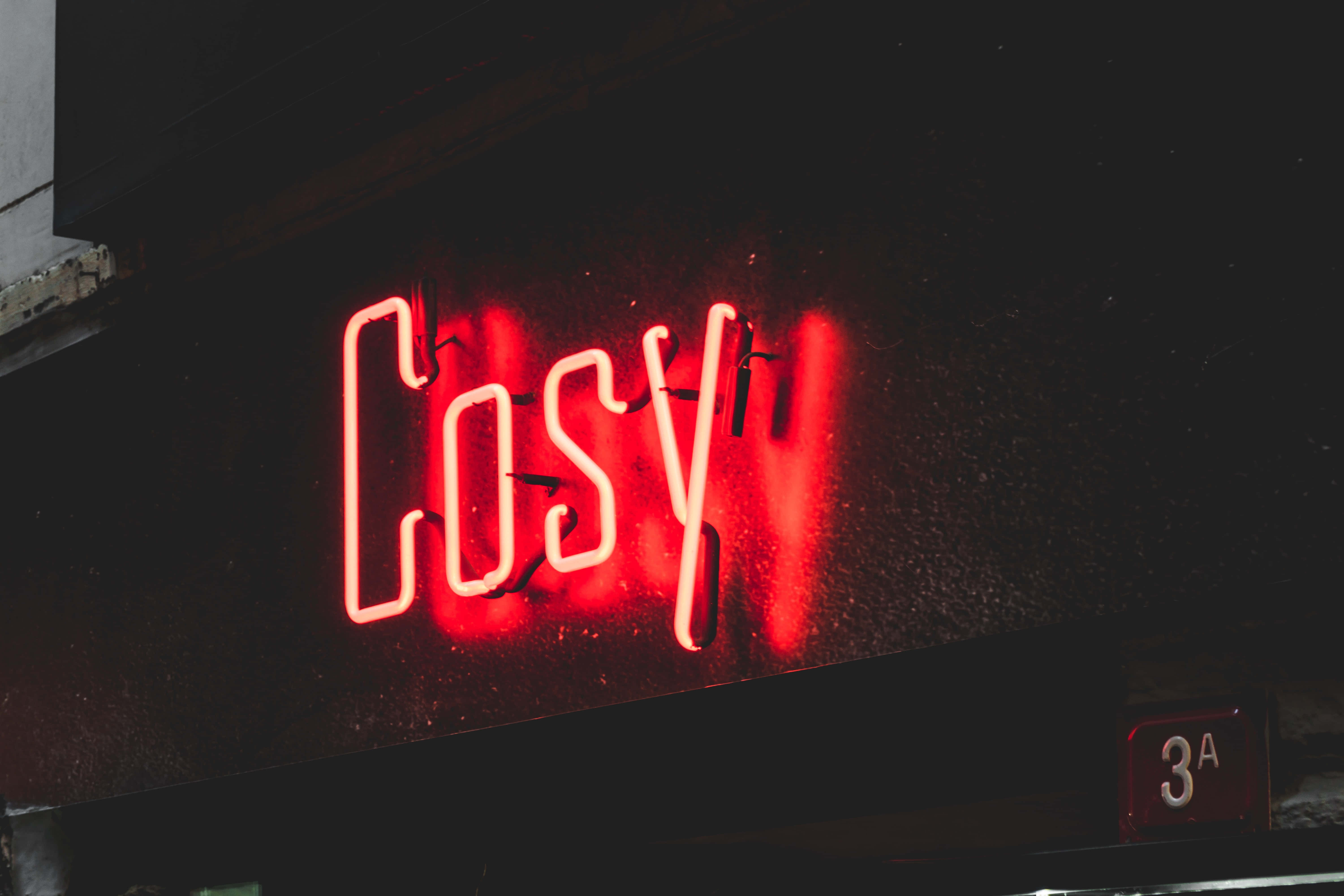 霓虹紅 桌布 壁紙 — 一個發光的紅色霓虹燈牌，上面寫著「Cosy」，在夜晚向黑暗潮濕的牆壁投射出溫暖的光芒。
