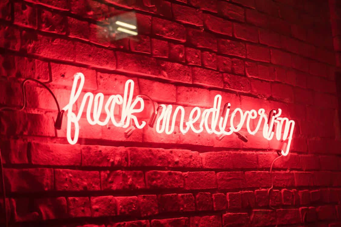 Red Neon 桌布 壁紙 — 一個發光的紅色霓虹燈牌，上面寫著「fuck mediocrity」，在昏暗的空間中將大膽的光芒投射在紋理磚牆上。