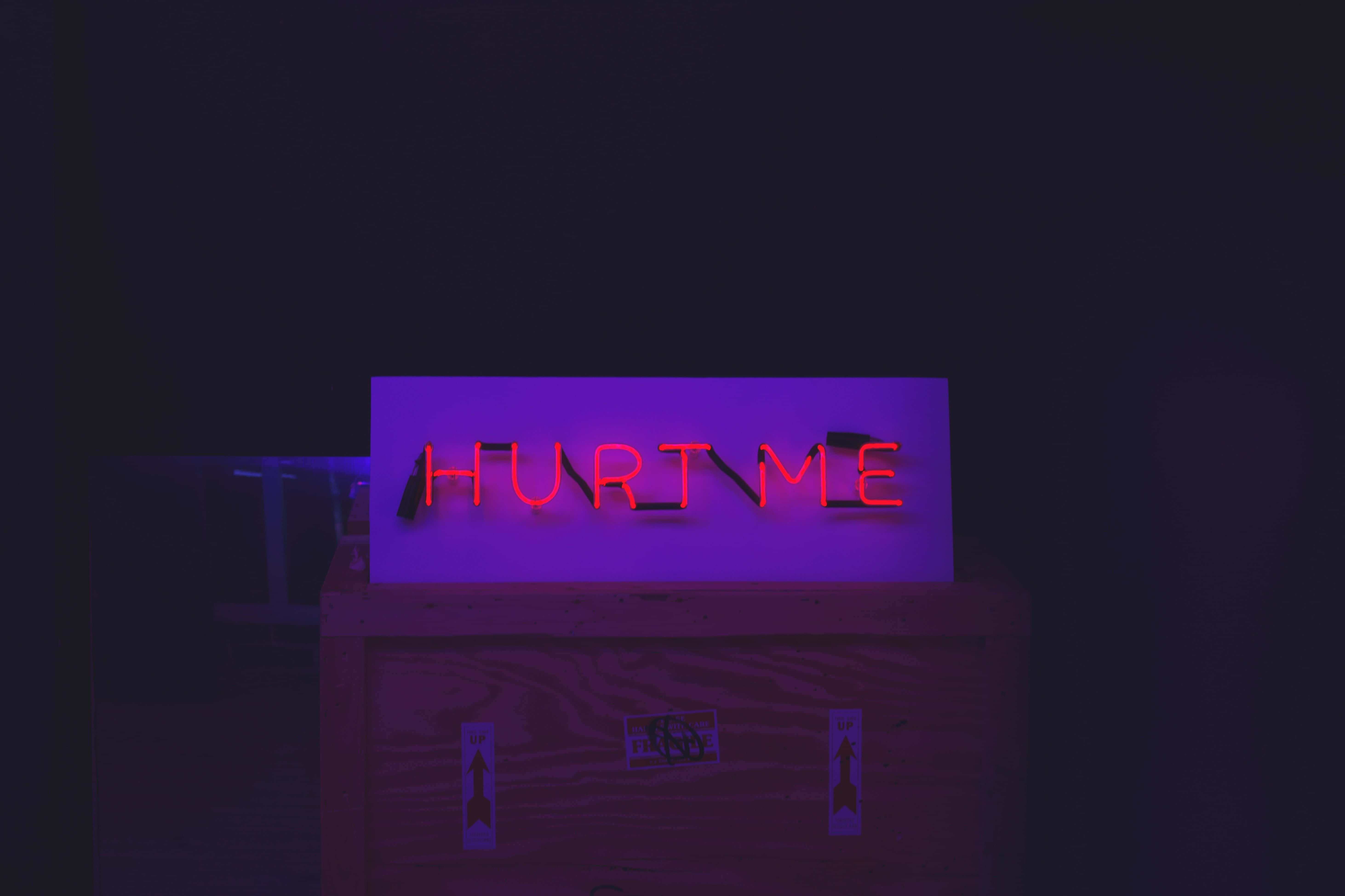 霓虹美學 桌布 壁紙 — 一個發光的紅色霓虹燈牌，上面寫著 'HURT ME'，在黑暗的房間中將紫色光暈投射在木箱上。
