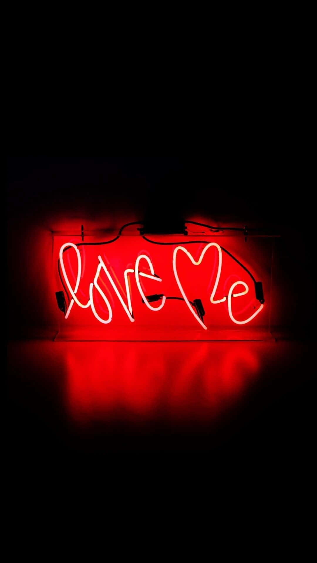 Neon Light 桌布 壁紙 — 一個發光的紅色霓虹燈牌，拼寫著 'Love Me'，其中 'o' 被一顆心取代，在黑暗的表面上投射出溫暖的倒影。