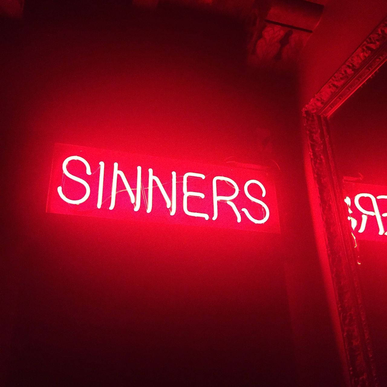 紅色動圖 桌布 壁紙 — 一個發光的紅色霓虹燈牌，拼寫著 SINNERS，在昏暗的室內環境中，將強烈的光芒投射在帶有華麗線條的深色牆壁上。