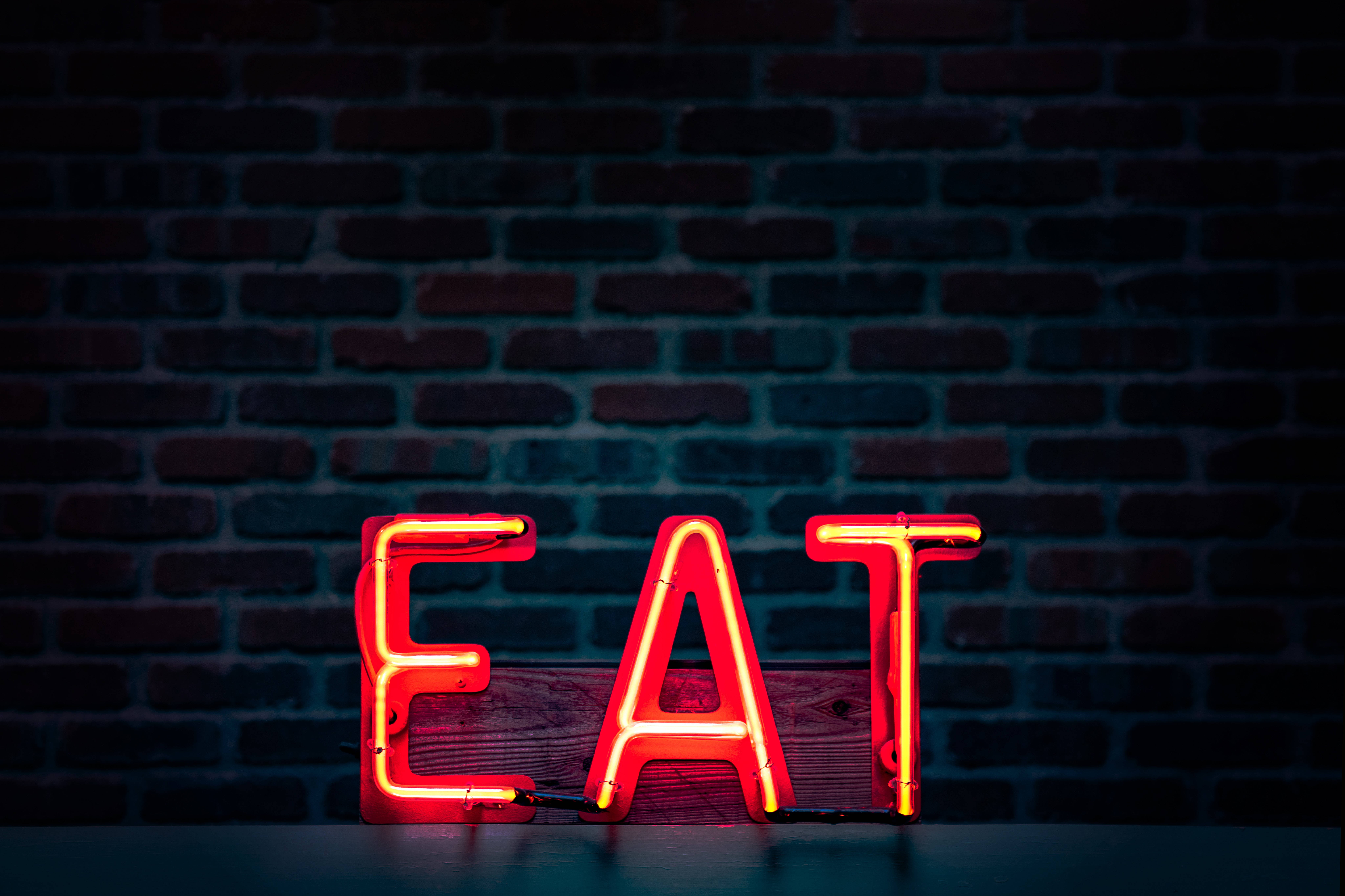 neon sign 桌布 壁紙 — 一個發光的紅色霓虹燈牌，拼寫著EAT，放在木質表面上，背景是深色磚牆。