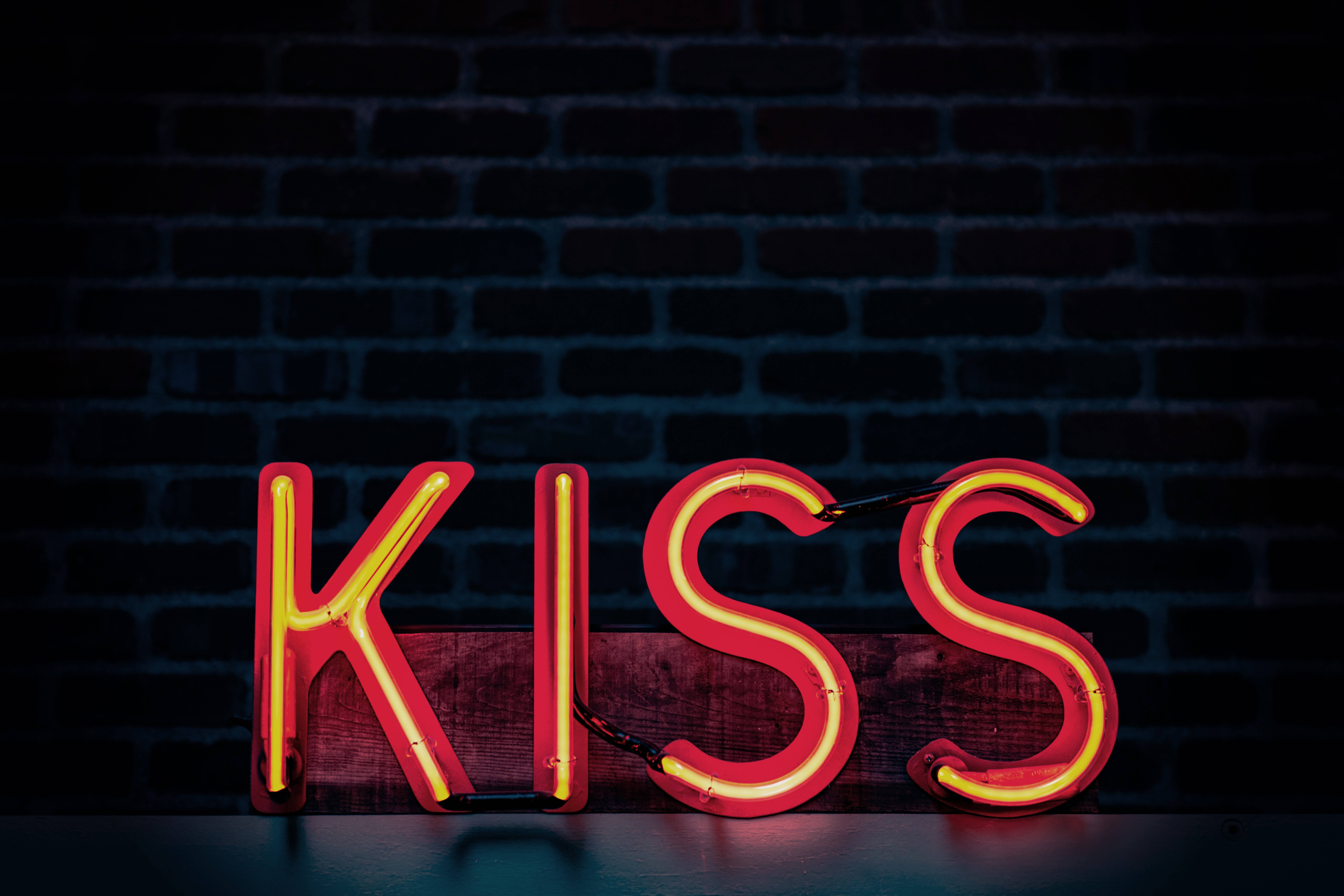 neon sign 桌布 壁紙 — 一個發光的紅色霓虹燈牌，拼寫著KISS，擱在木板上，靠著深色磚牆。