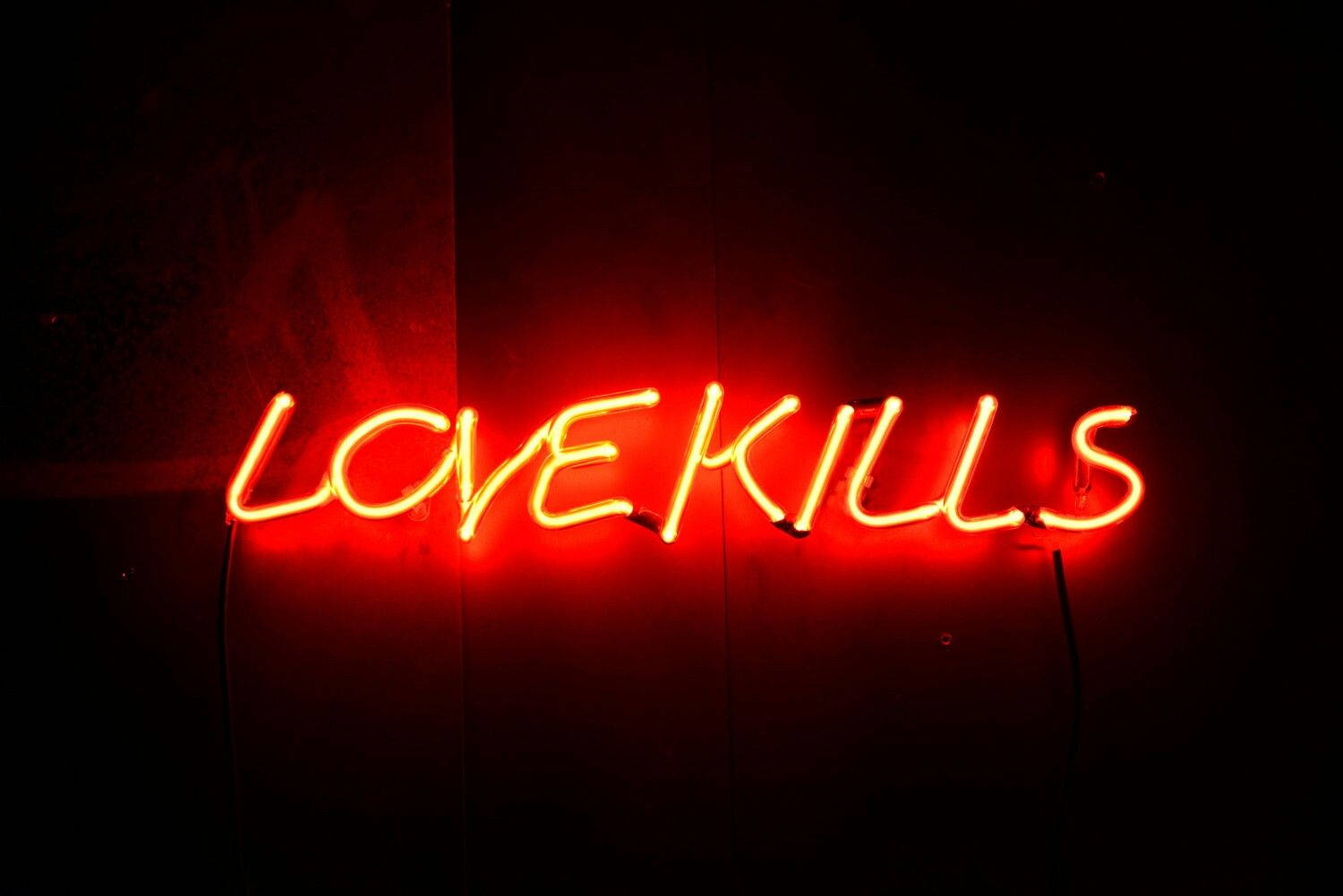 愛情語錄 桌布 壁紙 — 一個發光的紅色霓虹燈牌，拼寫著LOVEKILLS，在昏暗的空間中向深色紋理牆壁投射出溫暖的光芒。