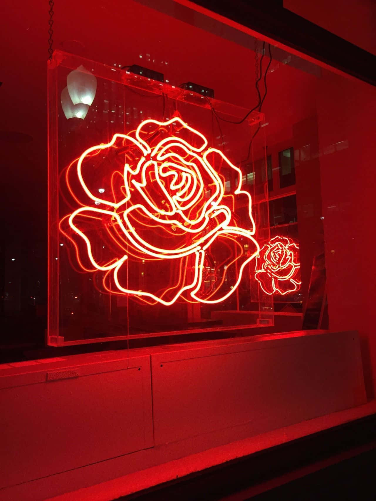 Neon Red Aesthetic 桌布 壁紙 — 一個發光的紅色霓虹玫瑰雕塑在玻璃箱中，在夜晚向黑暗的城市窗戶散發出溫暖的光芒。