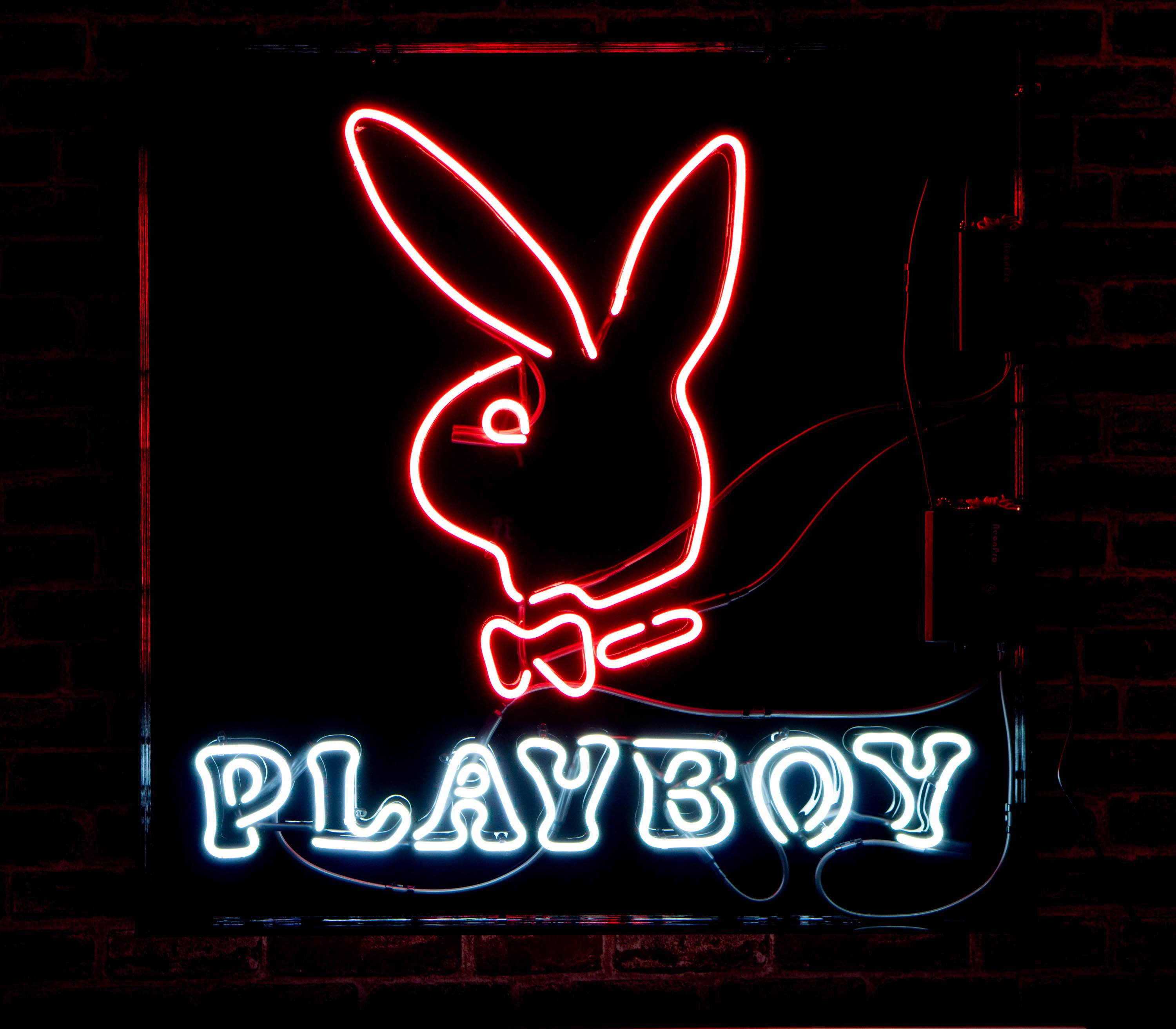 花花公子 桌布 壁紙 — 一個發光的紅藍色霓虹燈牌，上面有Playboy兔子標誌，背景是深色磚牆。