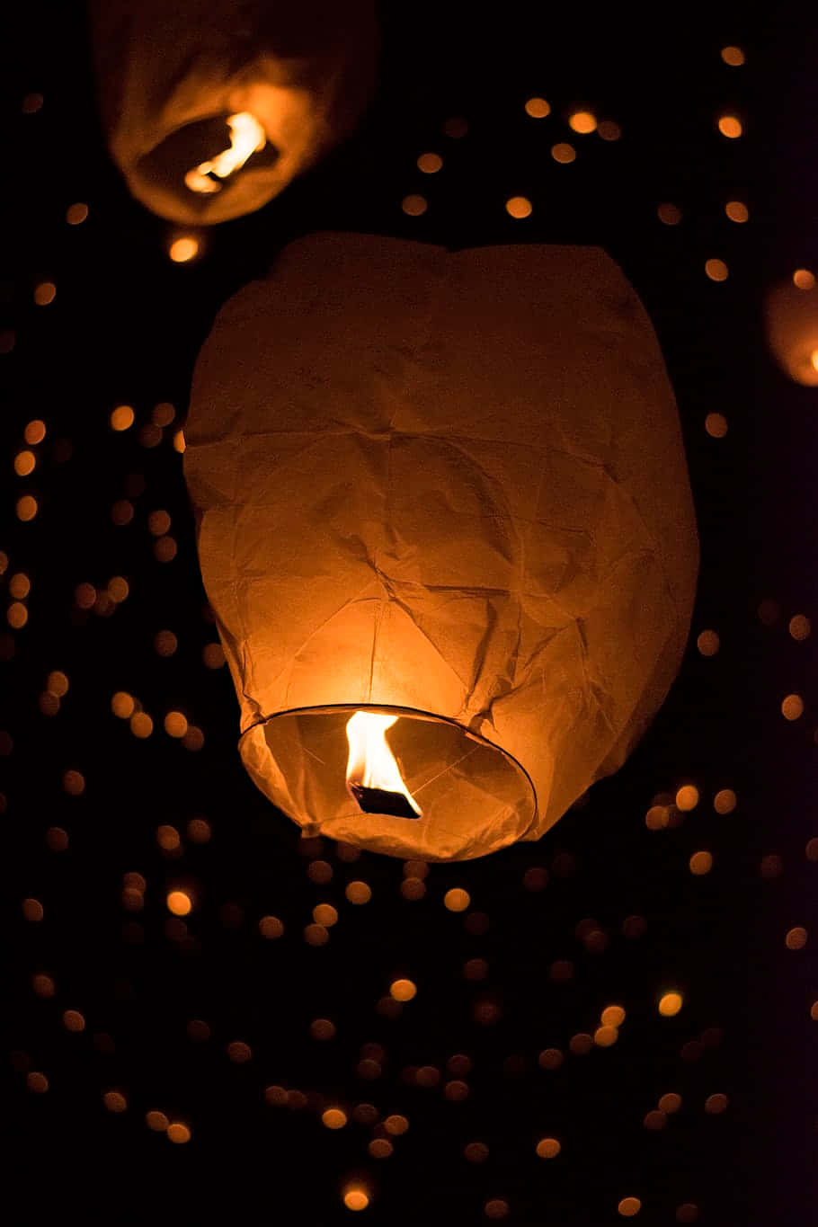 Lantern 桌布 壁紙 — 一個發光的紙燈籠在黑暗的天空中飄浮，天空點綴著來自其他燈籠的溫暖散景燈光。