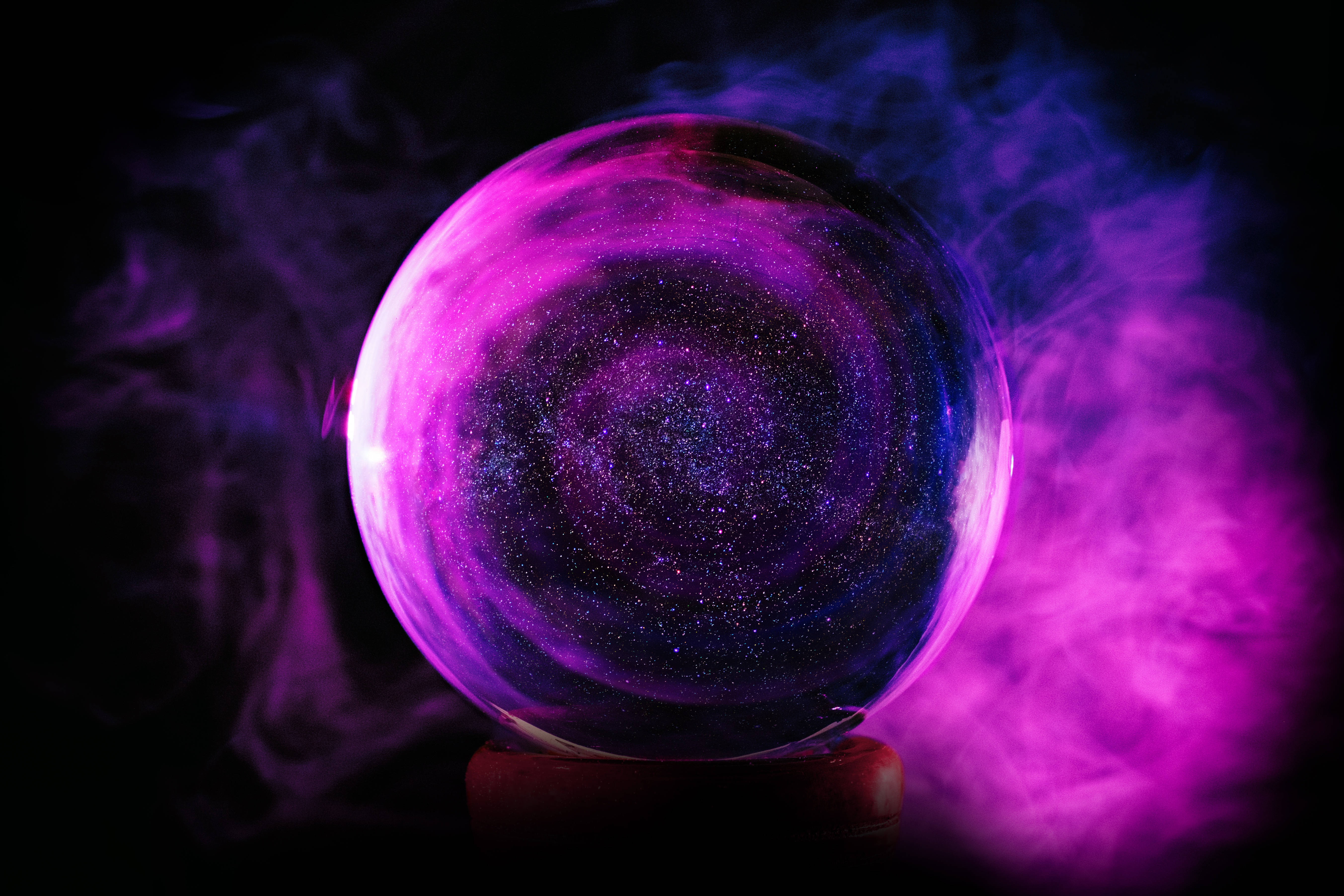 魔法動圖 桌布 壁紙 — 一個發光的紫色水晶球，充滿旋轉的宇宙星星，放在一個支架上，周圍環繞著空靈的煙霧，背景是黑暗的。