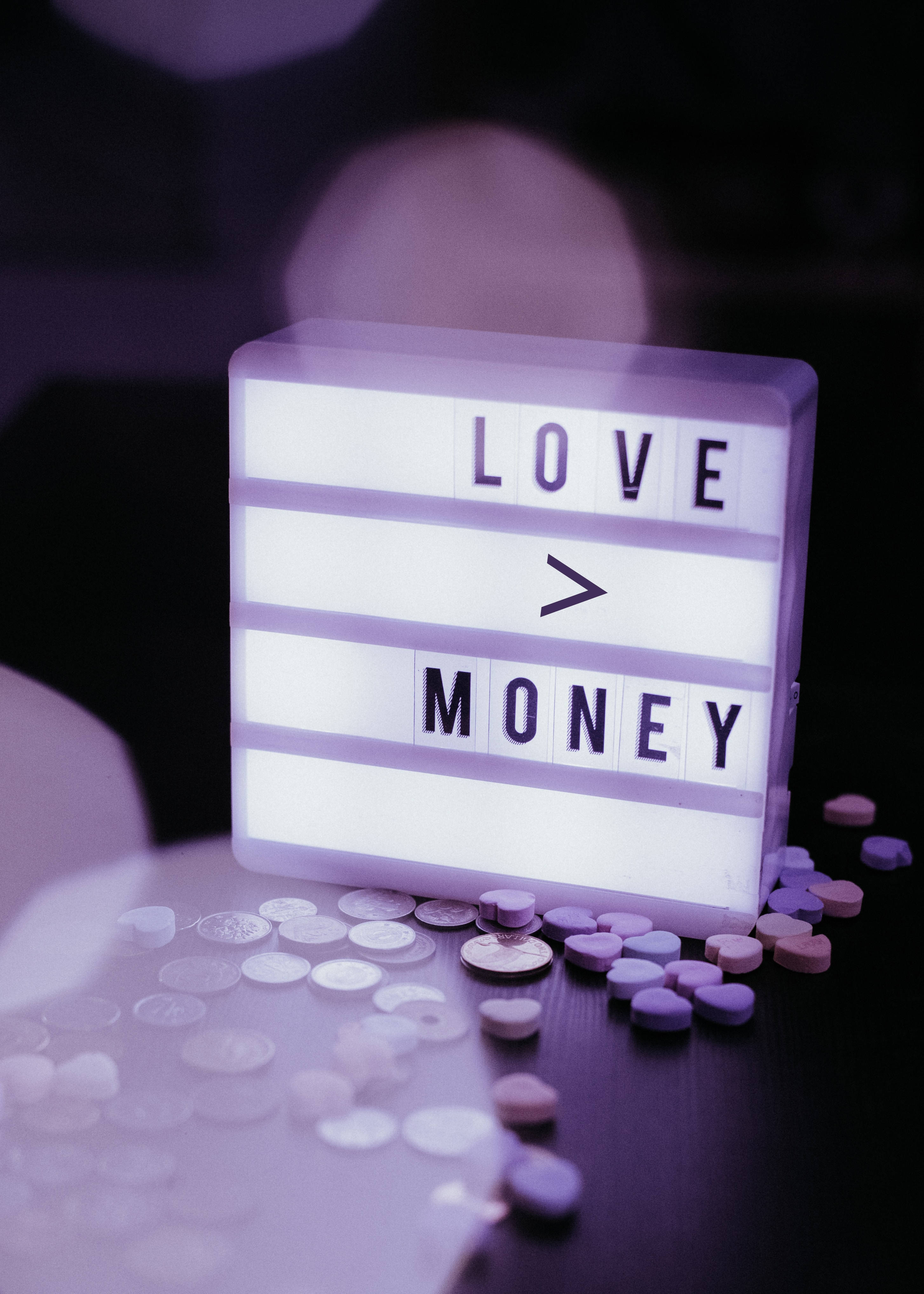 No Money 桌布 壁紙 — 一個發光的紫色燈箱顯示「LOVE > MONEY」，周圍散落著硬幣和藥丸在黑暗的表面上。