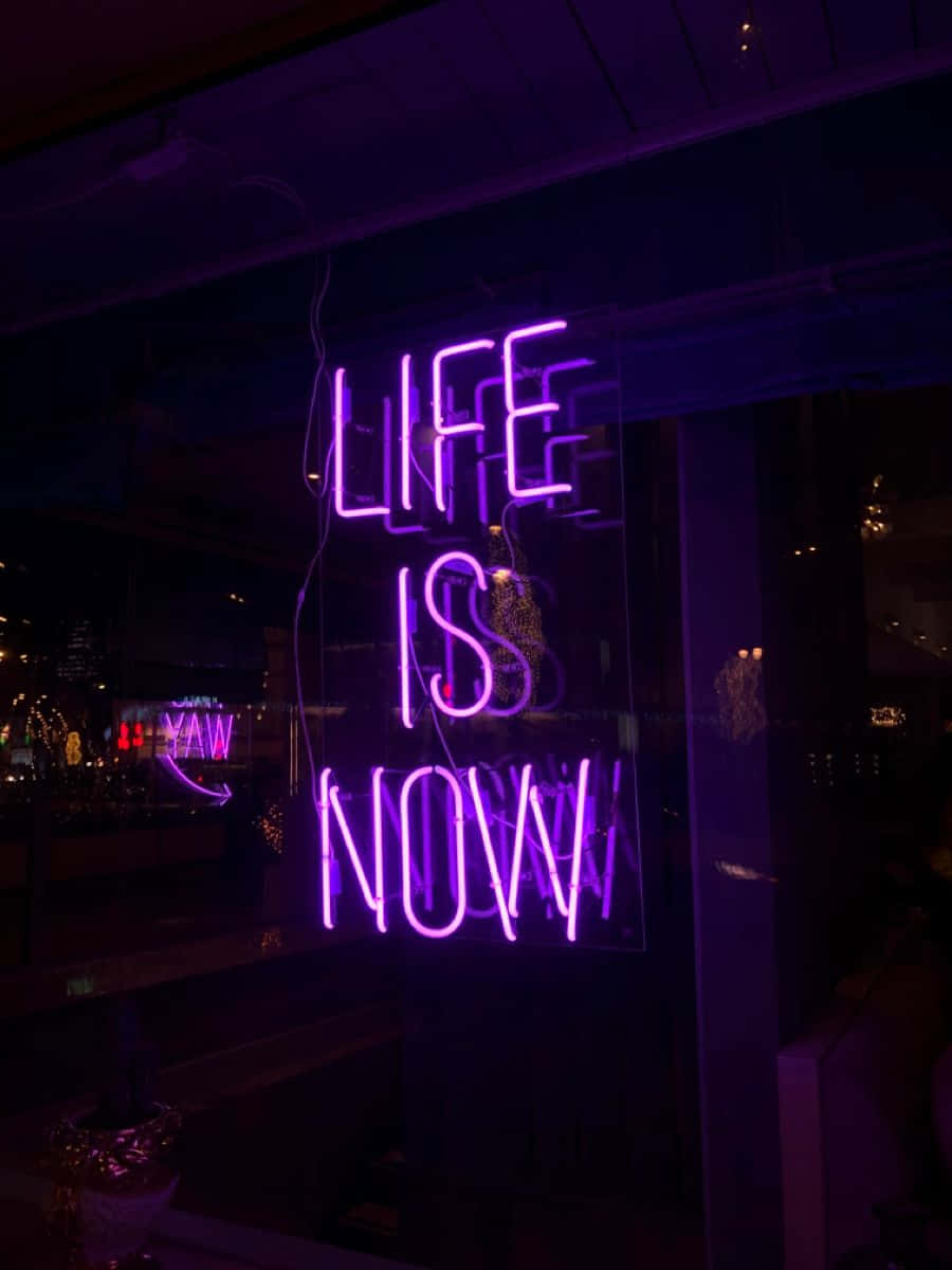 Neon Quotes 桌布 壁紙 — 一個發光的紫色霓虹燈牌，上面寫著「LIFE IS NOW」，在夜晚照亮了黑暗的城市窗戶。