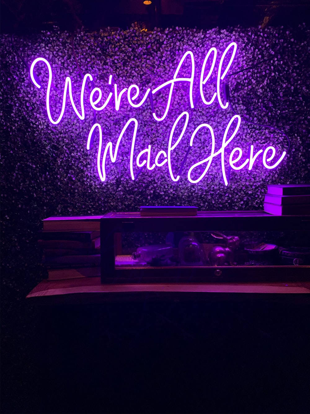 霓虹紫 iPhone 桌布 壁紙 — 一個發光的紫色霓虹燈牌，上面寫著 We're All Mad Here，照亮了一個黑暗的房間，背景是帶紋理的牆壁，下方堆疊著書籍。