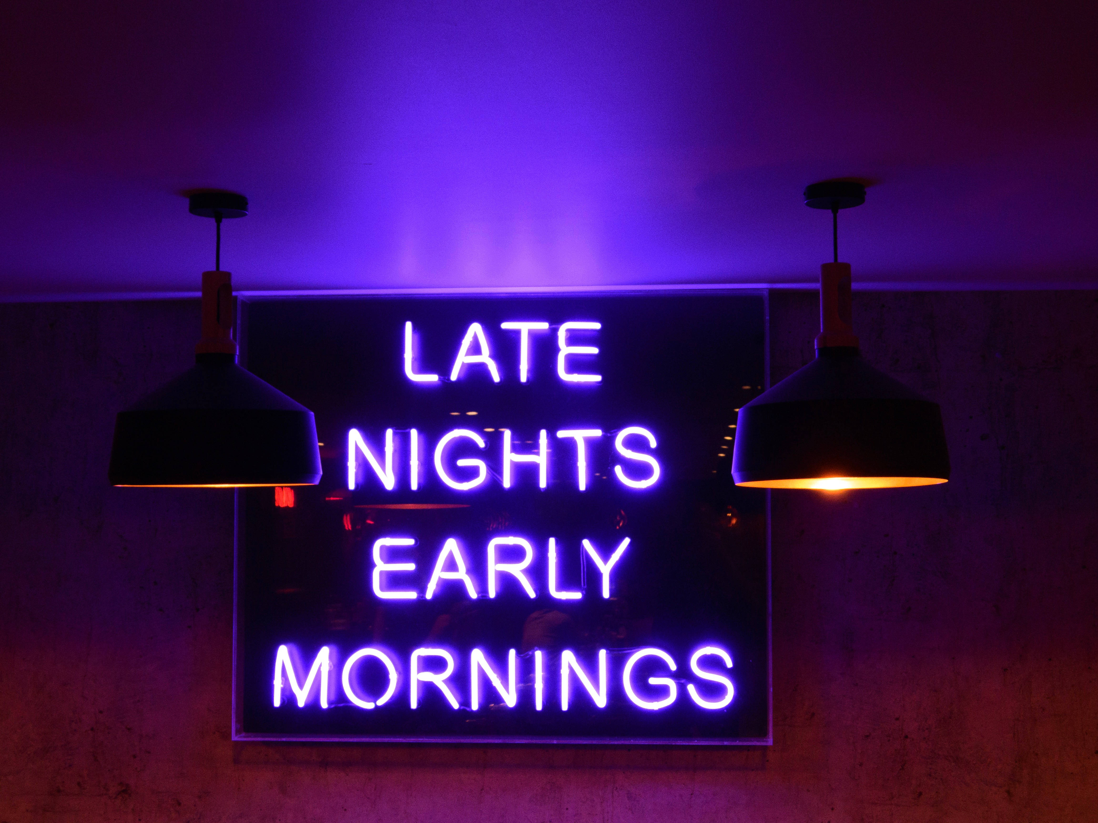 4K 霓虹 iPhone 桌布 壁紙 — 一個發光的紫色霓虹燈牌，上面寫著「late nights early mornings」，懸掛在兩盞吊燈之間，房間光線昏暗。