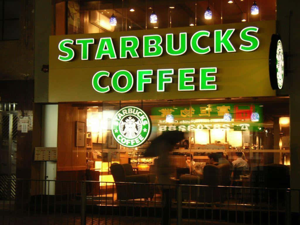 星巴克 桌布 壁紙 — 一個發光的綠色Starbucks標誌照亮了夜晚繁忙的都市咖啡店，裡面有模糊的顧客。