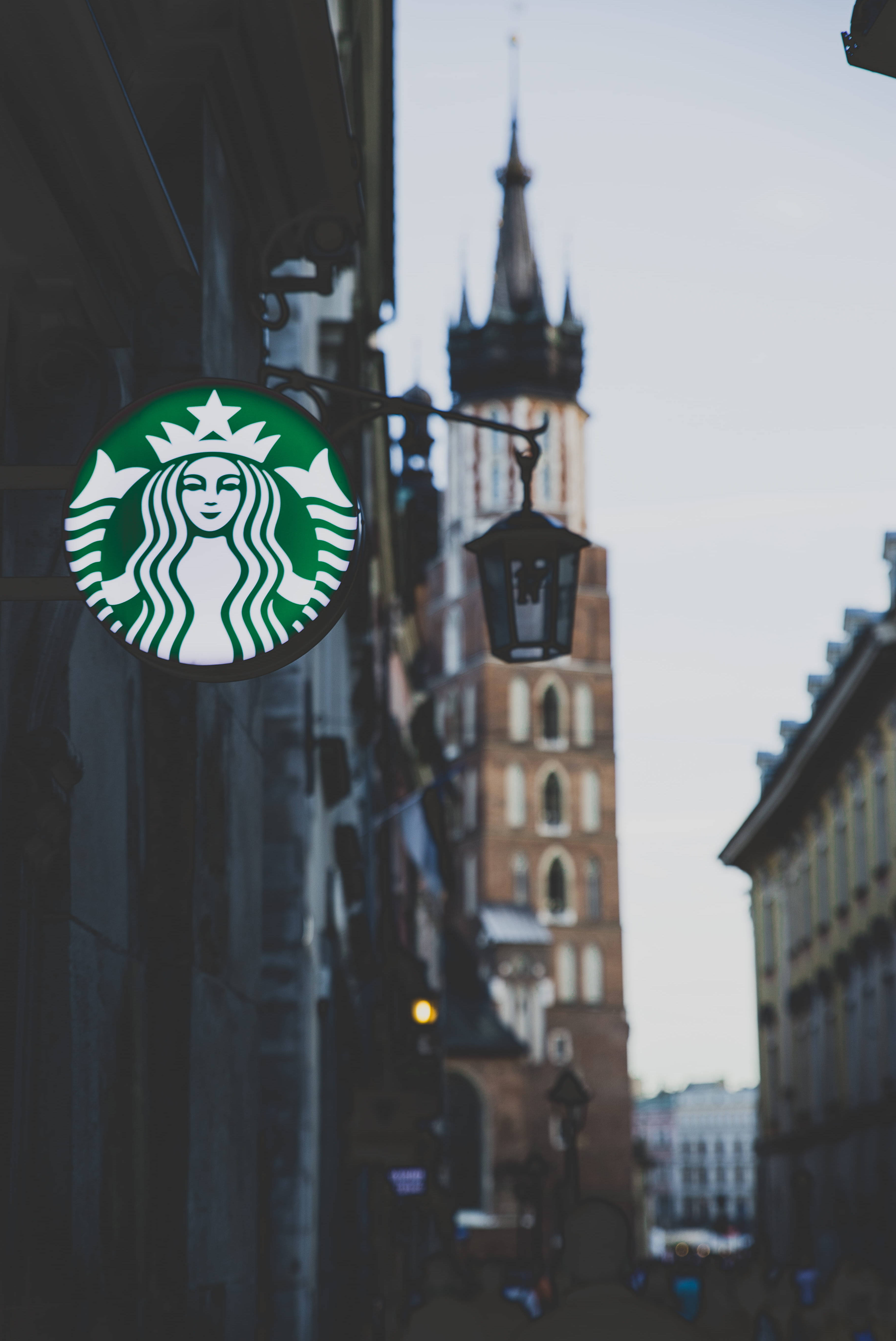 星巴克 桌布 壁紙 — 一個發光的綠色Starbucks標誌掛在一棟建築物上，背景模糊中可見一座歷史塔樓。