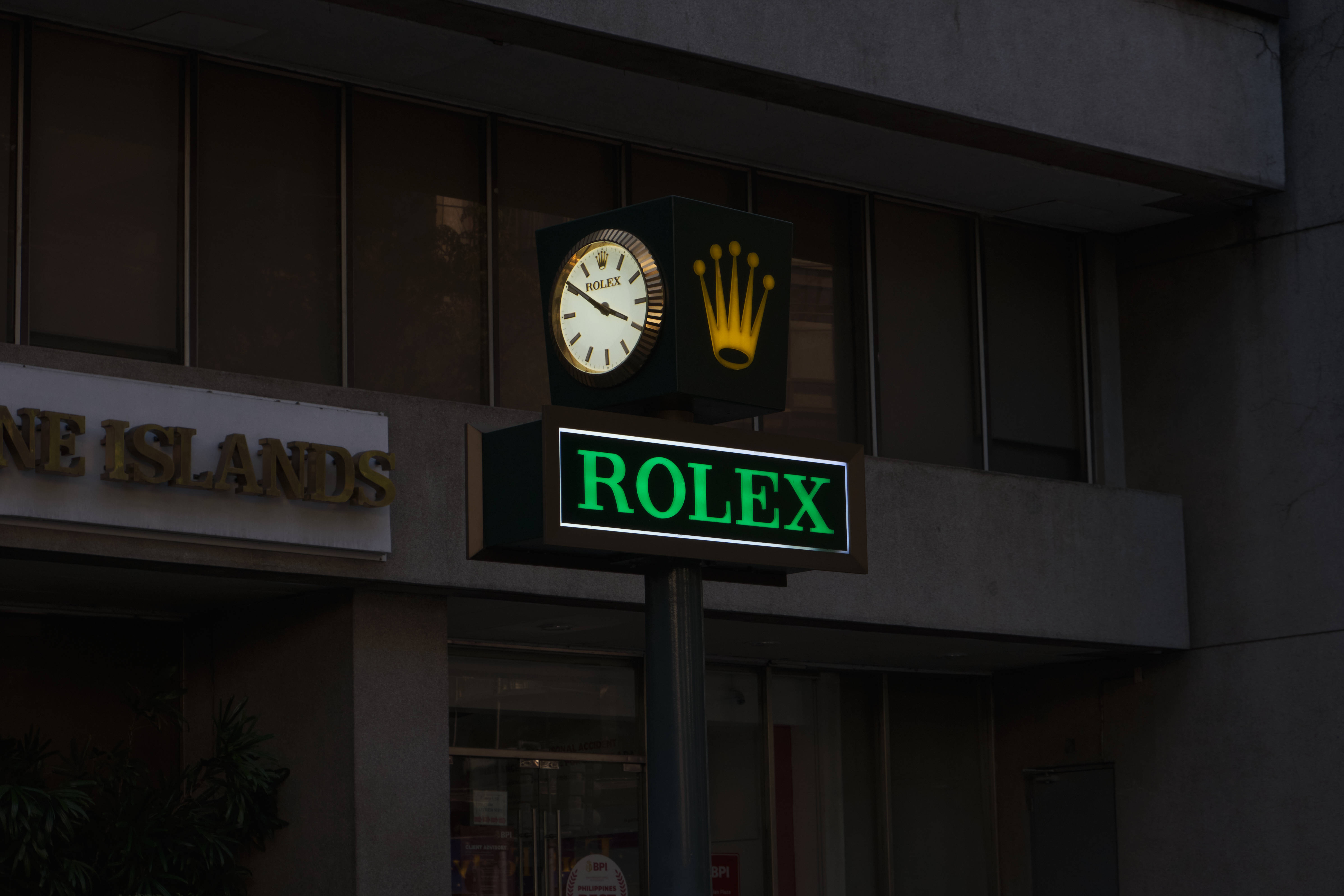 Rolex 桌布 壁紙 — 一個發光的綠色勞力士標誌和時鐘安裝在黃昏時分的建築物外，帶有深色窗戶和混凝土外牆。