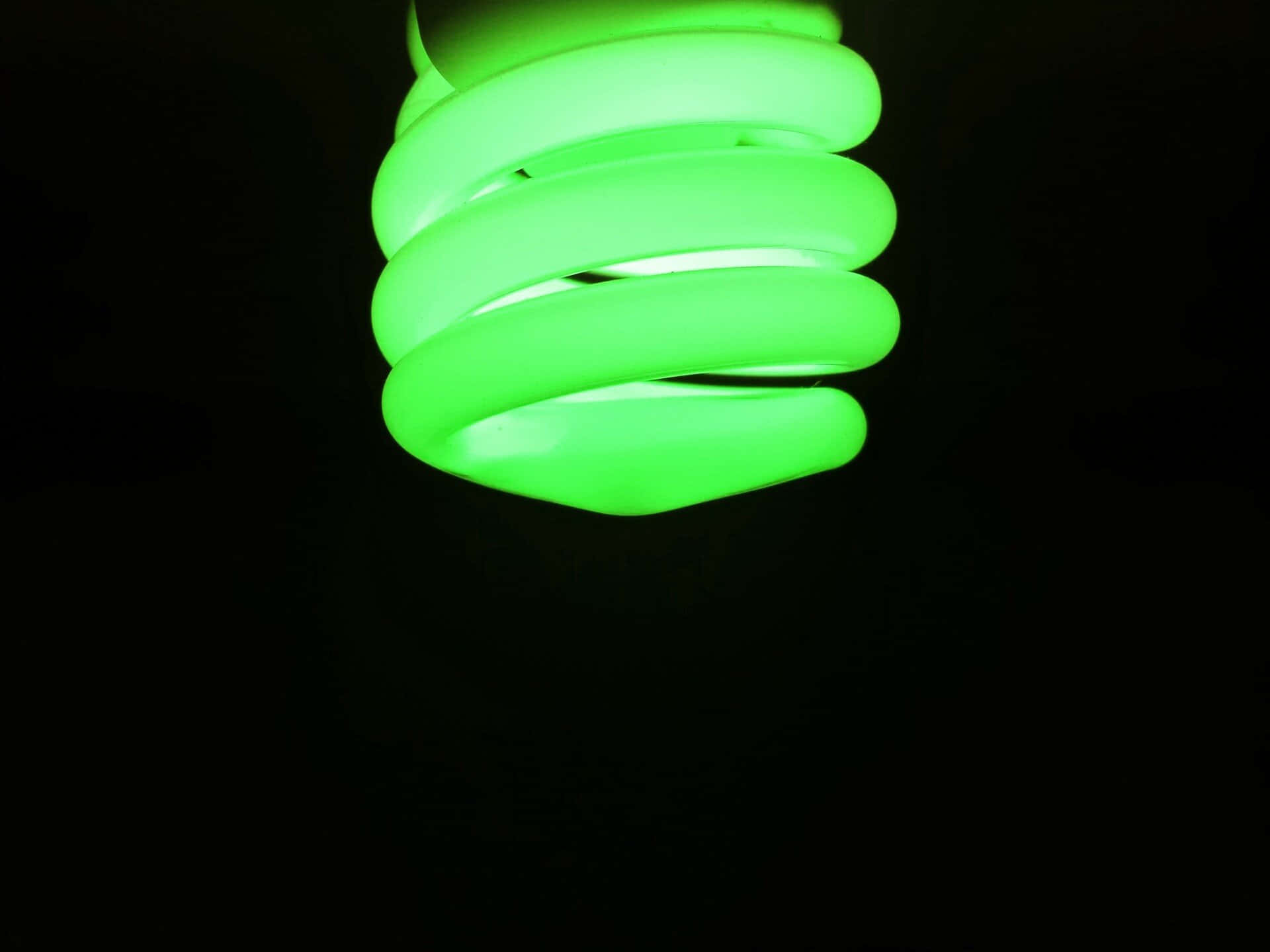 Green Led 桌布 壁紙 — 一個發光的綠色緊湊型螢光燈泡在漆黑背景上盤旋，以極簡構圖投射柔和光線。