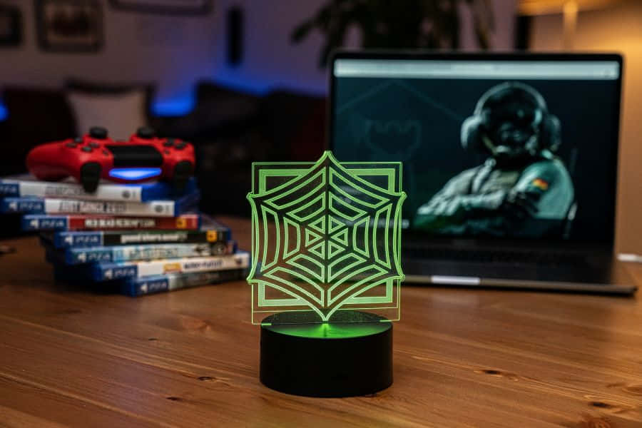 Rainbow Six Siege Jager 桌布 壁紙 — 一個發光的綠色蜘蛛網燈放在木桌上，旁邊有一個紅色PS5手把和一台顯示遊戲玩家的筆記型電腦。