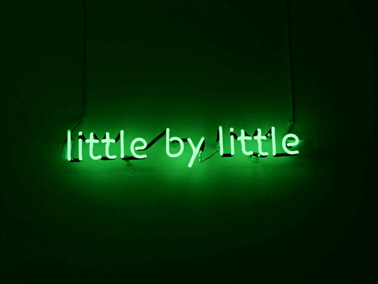 lights 桌布 壁紙 — 一個發光的綠色霓虹燈牌，上面寫著 little by little，掛在黑暗的牆上，投射出柔和的環境光。