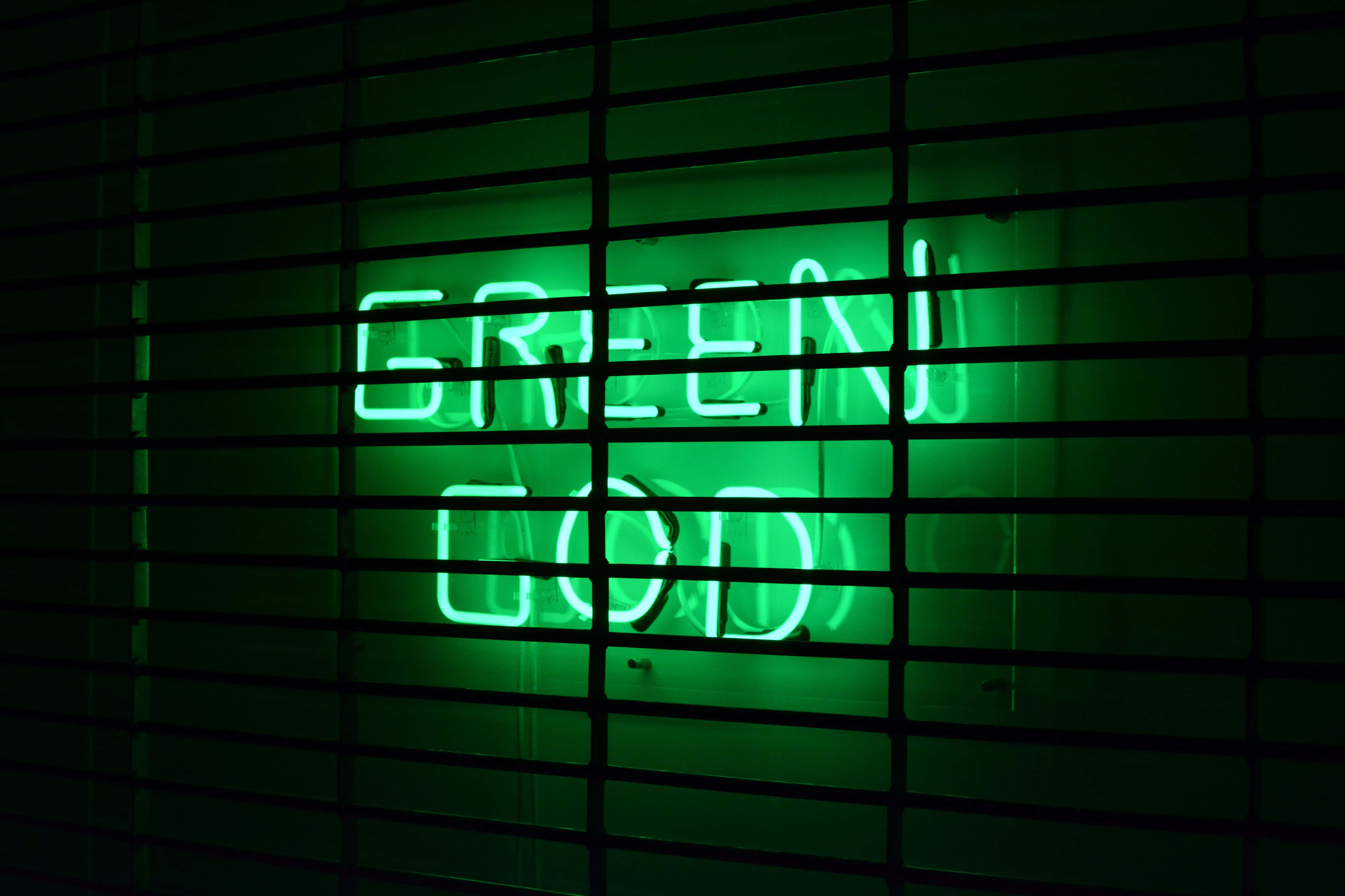 桌布 壁紙 — 一個發光的綠色霓虹燈牌，上面寫著GREEN GOD，在昏暗的房間中透過深色橫向百葉窗照射出來。