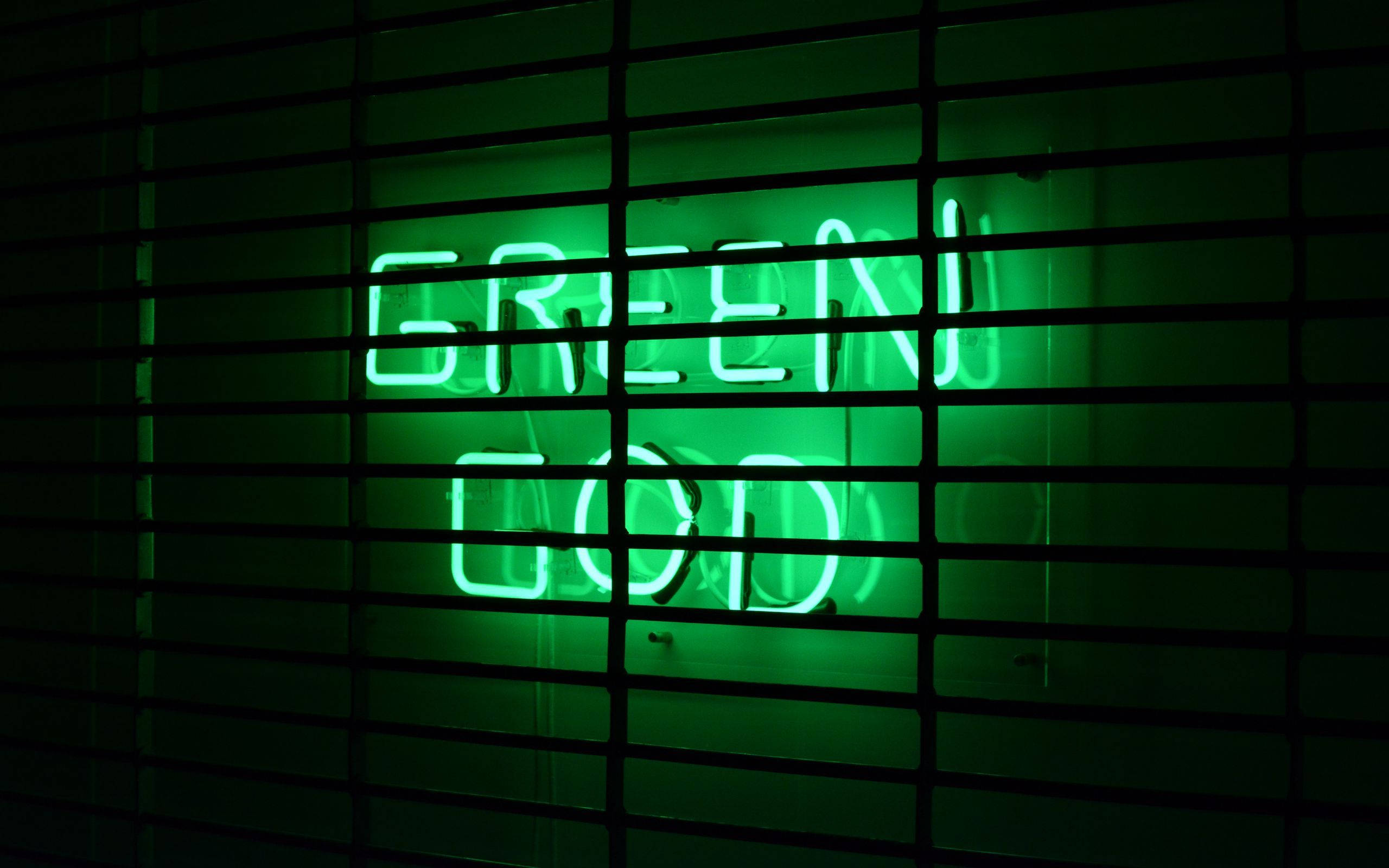 霓虹綠美學 桌布 壁紙 — 一個發光的綠色霓虹燈牌，上面寫著 GREEN GOD，透過昏暗房間的深色橫向百葉窗照射出來。