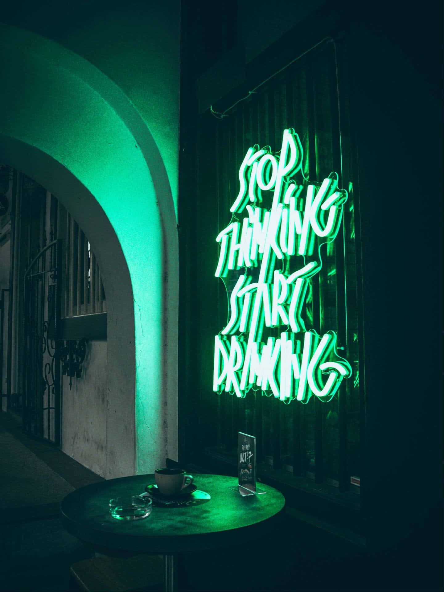 Green Led 桌布 壁紙 — 一個發光的綠色霓虹燈牌，上面寫著 stop thinking start drinking，照亮了一條昏暗的小巷，旁邊有一張小桌子和一個咖啡杯。