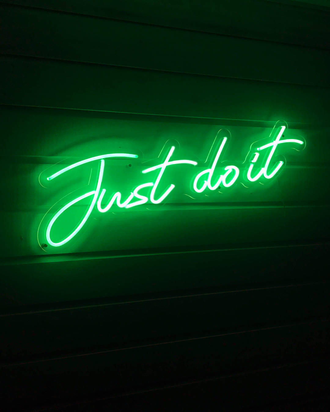 綠色美學 桌布 壁紙 — 一個發光的綠色霓虹燈牌，以草書寫著 'Just do it'，照亮了深色木牆。