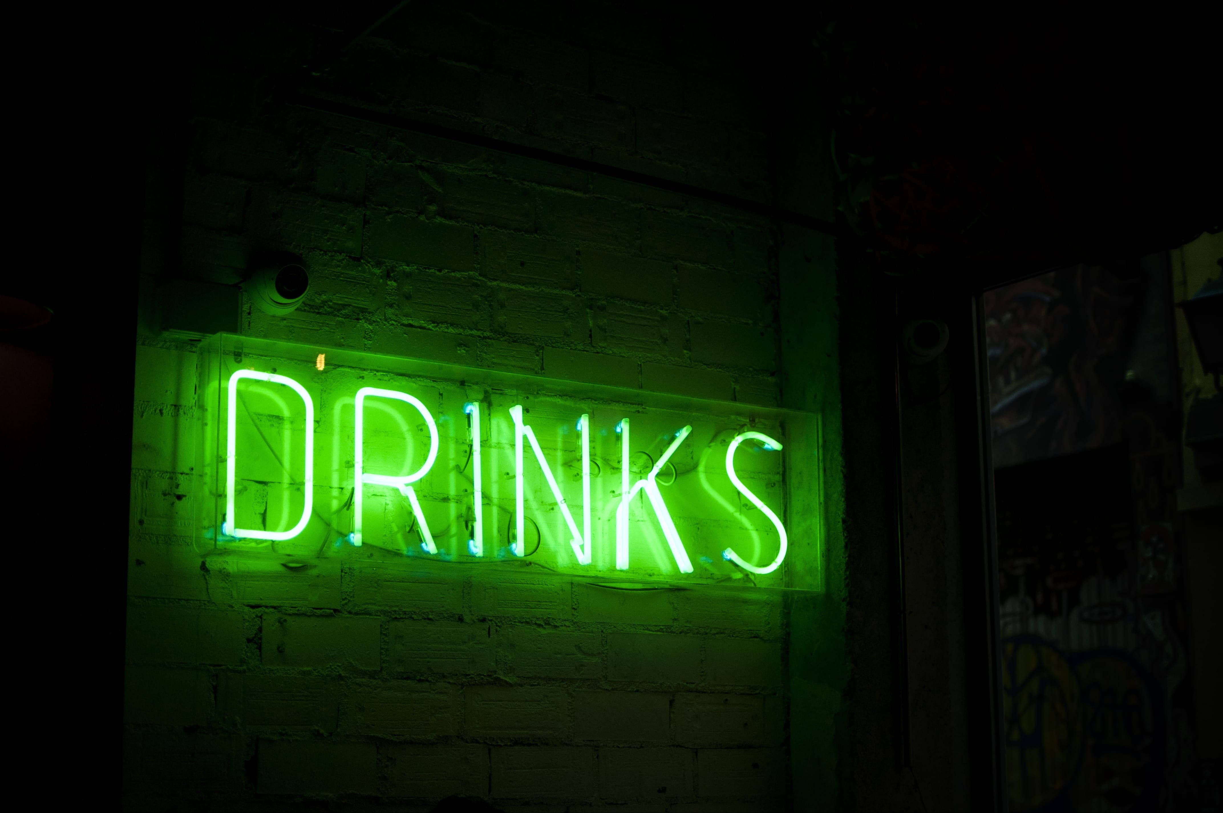 螢光綠 桌布 壁紙 — 一個發光的綠色霓虹燈牌，拼寫著DRINKS，在昏暗的城市小巷中，將電光般的光芒投射在粗糙的磚牆上。