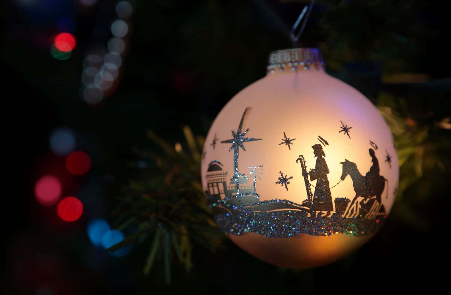Religious Christmas 桌布 壁紙 — 一個發光的聖誕裝飾品，上面有耶穌誕生場景的剪影，掛在帶有彩色散景燈光的樹上。