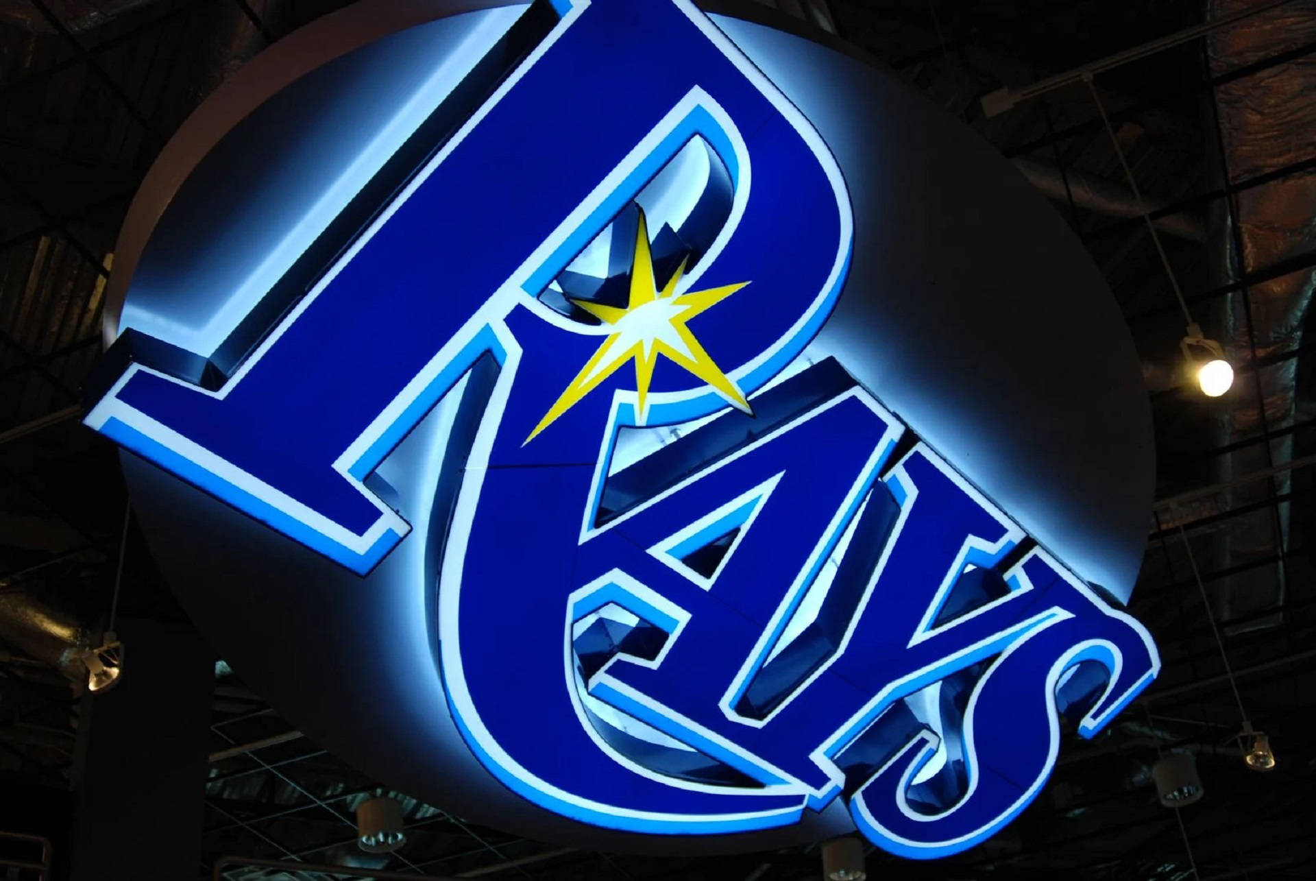 Rays 桌布 壁紙 — 一個發光的藍色Rays標誌，帶有一顆黃色星星，在黑暗的天花板下，有裸露的橫樑和吊燈，明亮地閃耀。