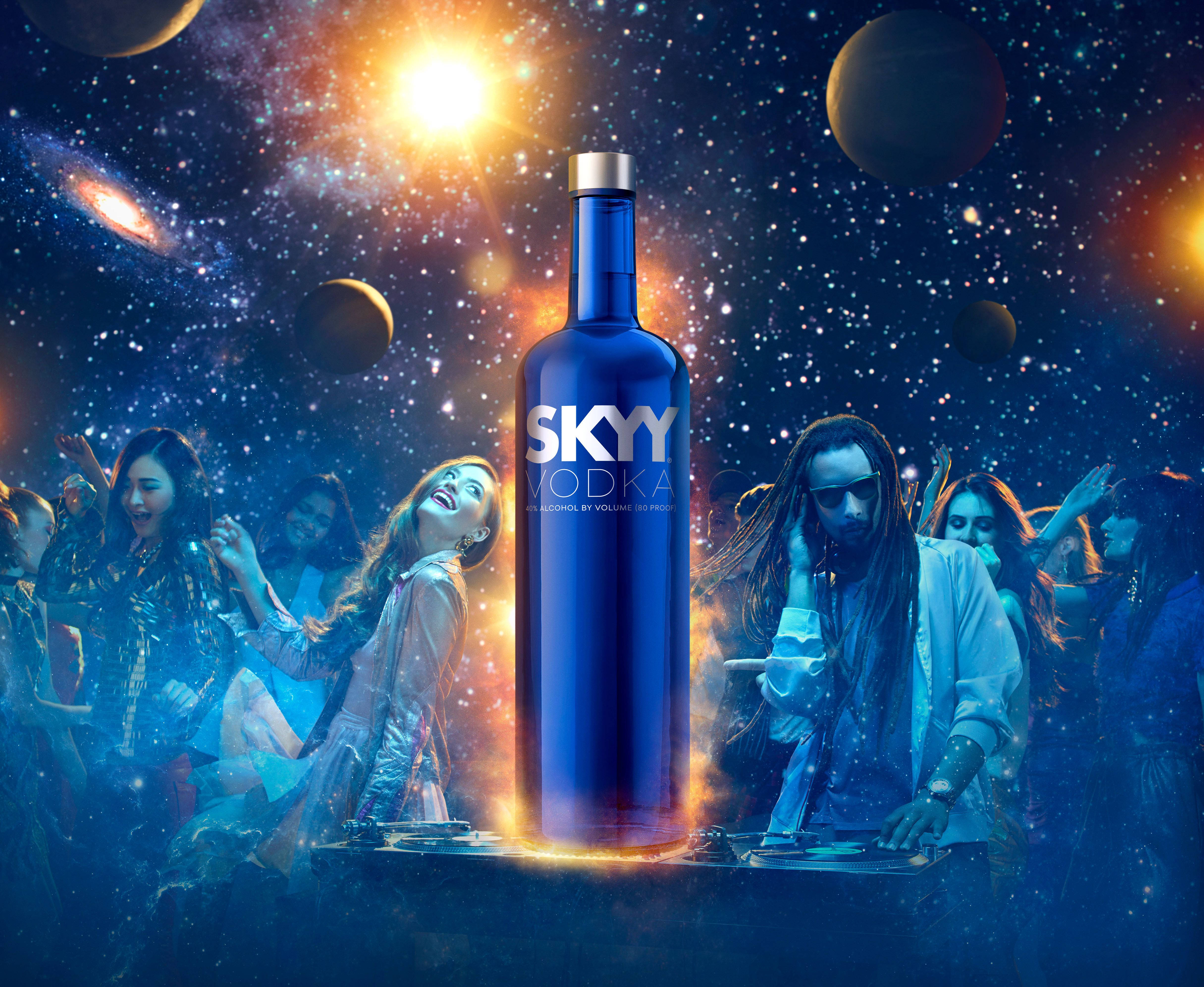 Drink 桌布 壁紙 — 一個發光的藍色Skyy Vodka酒瓶站在舞台中央，DJ和派對參與者在充滿星星、行星和星雲的充滿活力的宇宙天空下跳舞。