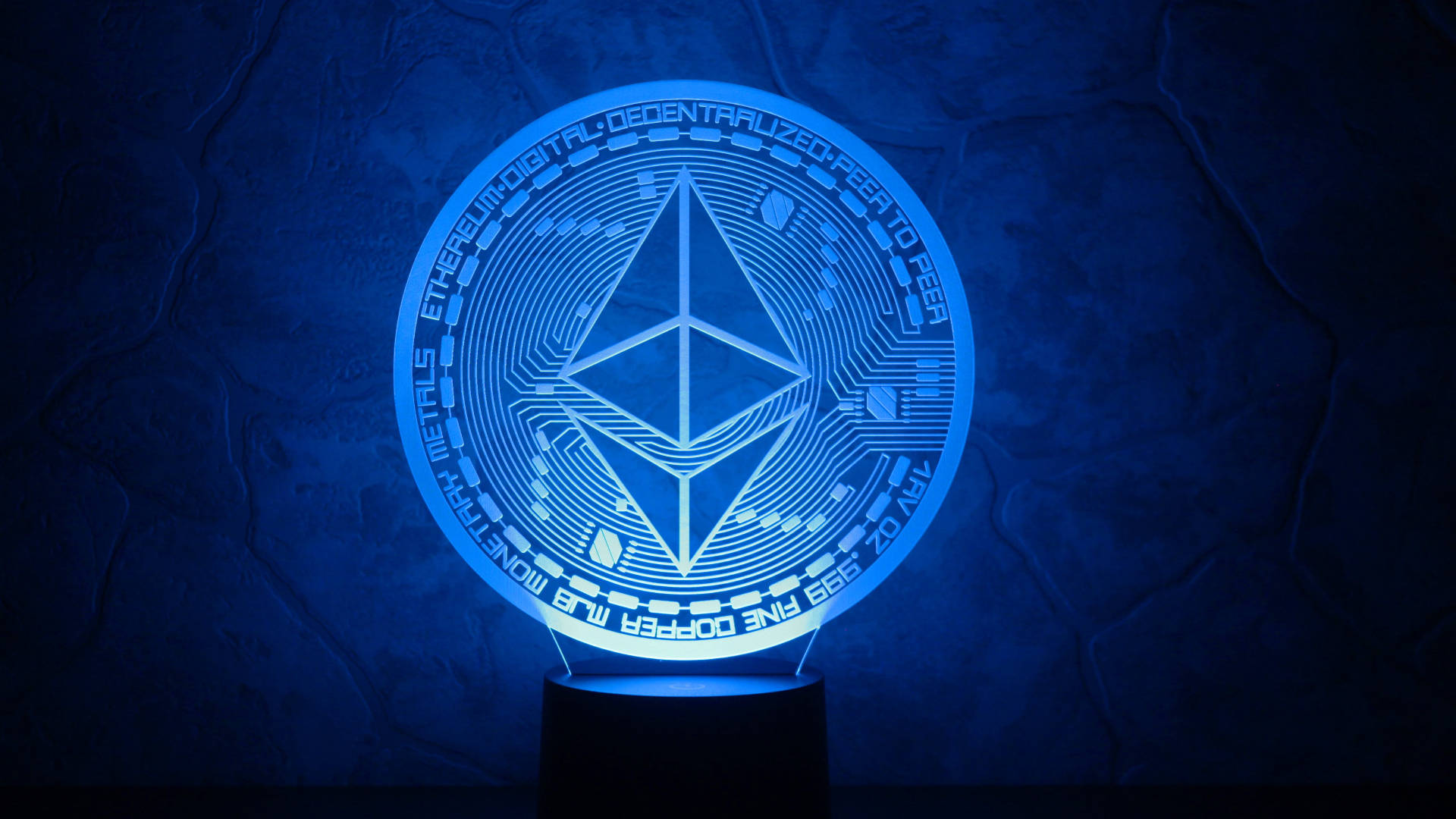 以太坊 桌布 壁紙 — 一個發光的藍色以太坊加密貨幣燈，帶有電路設計，在深色紋理牆壁上照亮。