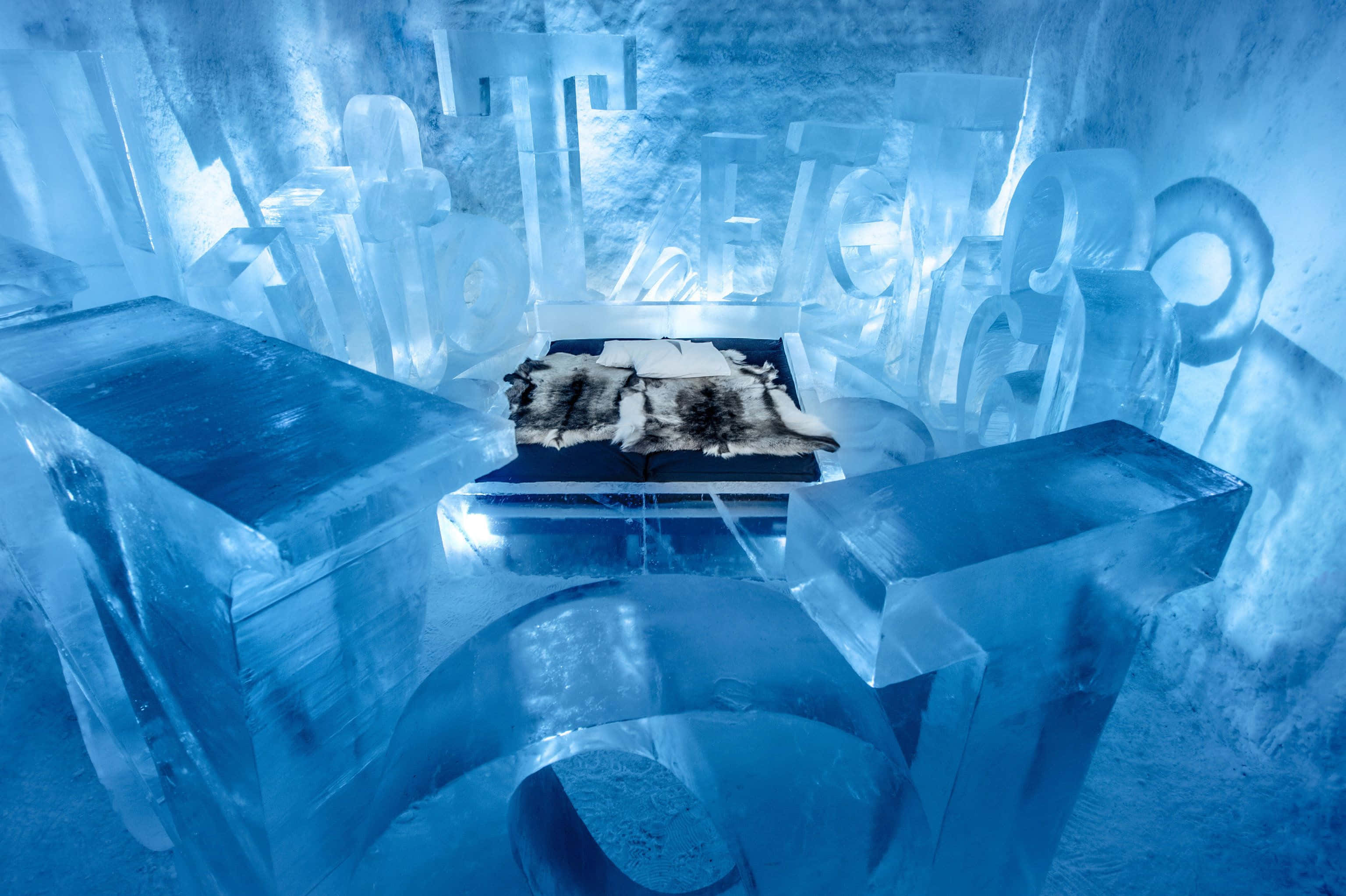 Ice Hotel 桌布 壁紙 — 一個發光的藍色冰室，配有雕刻家具和毛皮地毯，在冷色調人造燈光下。