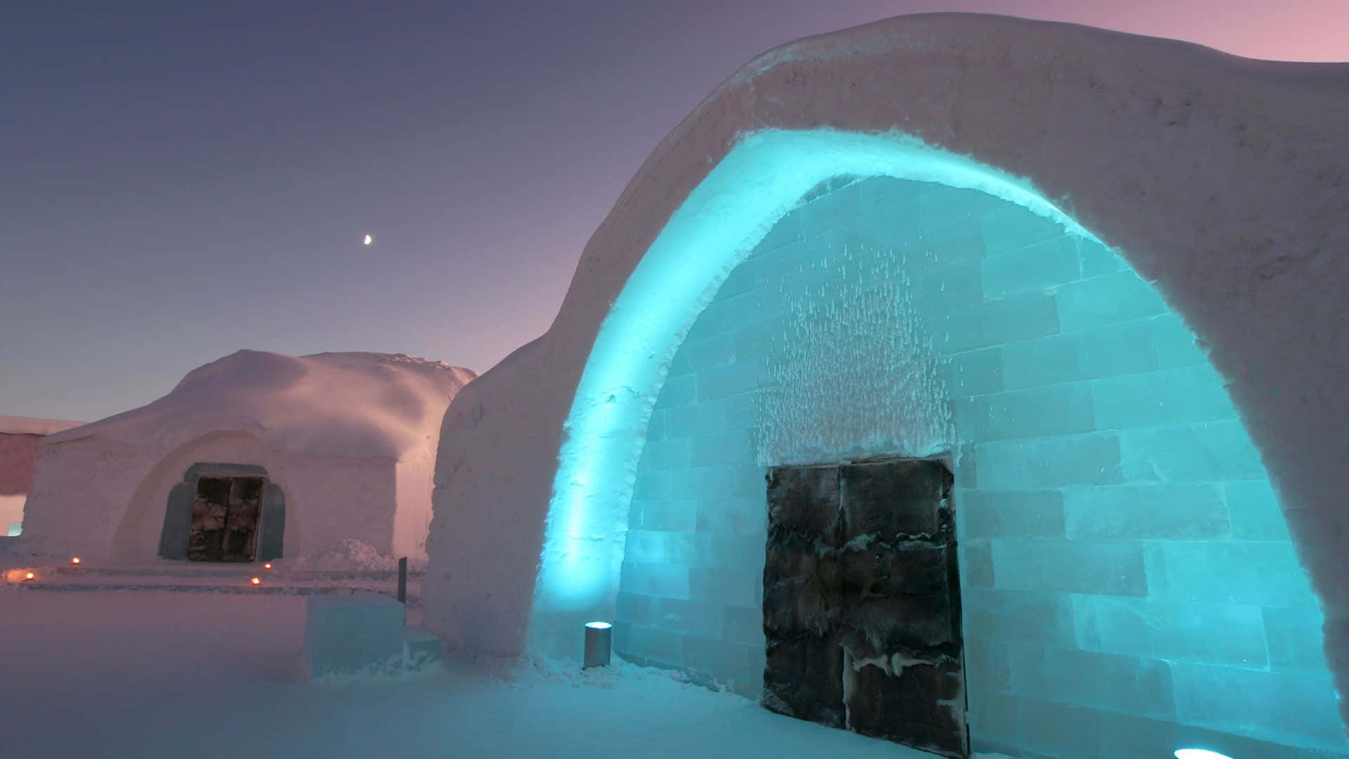 Ice Hotel 桌布 壁紙 — 一個發光的藍色冰屋，帶有深色木門，矗立在星空下的雪黃昏中。