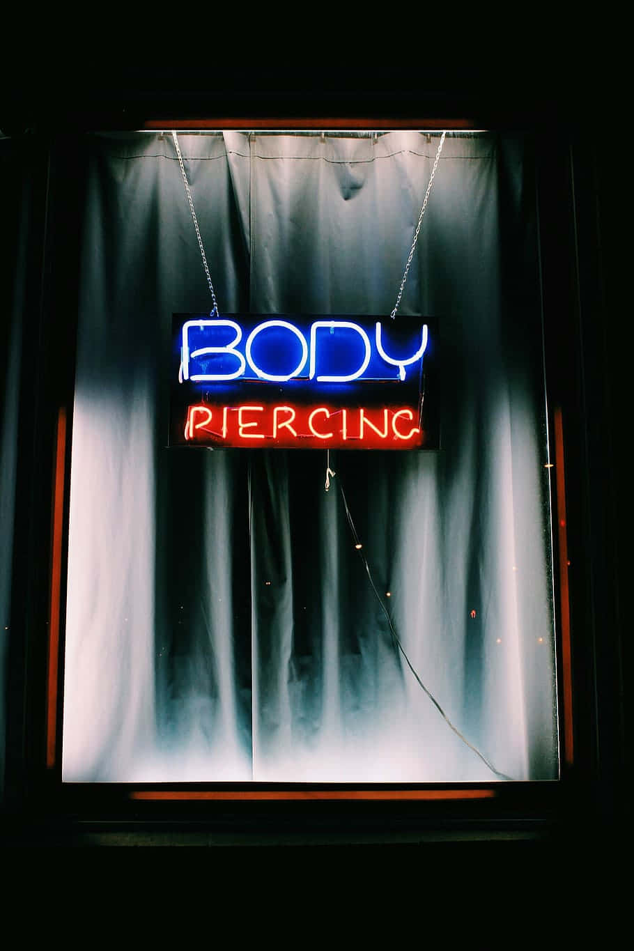 curtain opening 桌布 壁紙 — 一個發光的藍色和紅色霓虹燈招牌，上面寫著「BODY PIERCING」，掛在一個由深色窗簾框住的昏暗窗戶中。