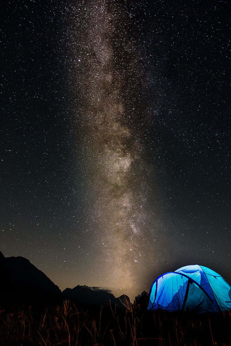 老友記 桌布 壁紙 — 一個發光的藍色帳篷坐落在令人嘆為觀止的星空下，銀河垂直延伸在黑暗的山脈之上。