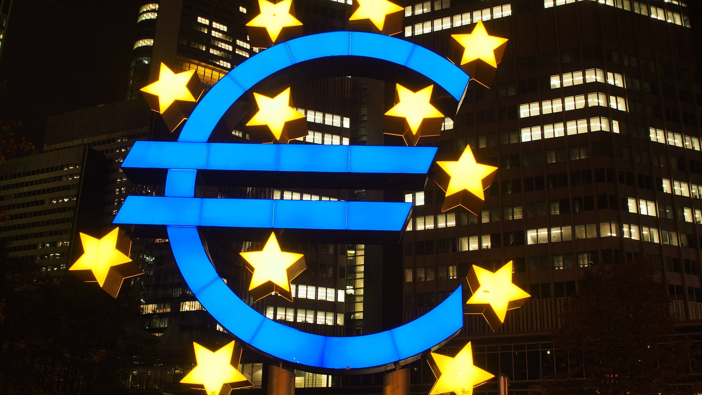 Europe 桌布 壁紙 — 一個發光的藍色歐元符號，被金色星星環繞，在夜晚點亮的摩天大樓背景下閃耀。