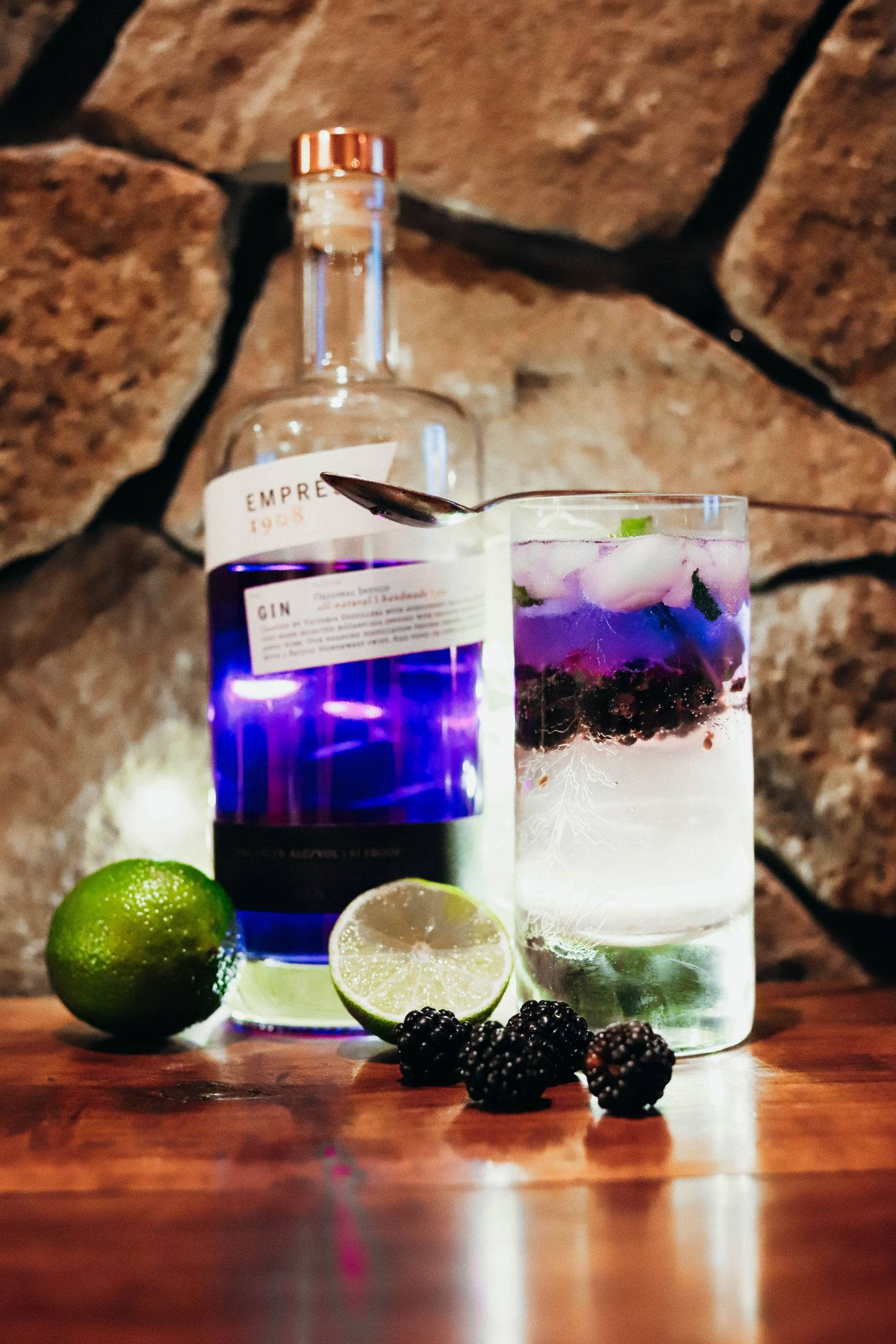cocktail 桌布 壁紙 — 一個發光的藍色琴酒瓶和一個分層的雞尾酒，搭配黑莓和萊姆，放在木桌上，背景是質樸的石牆。