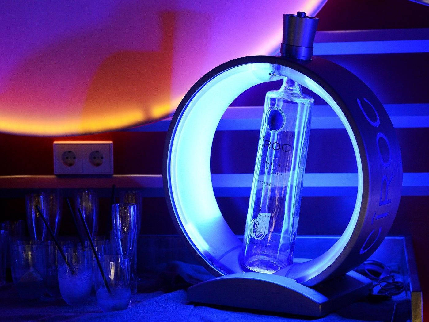 Drink 桌布 壁紙 — 一個發光的藍色環形燈照亮了一個標有 'Ciroc' 品牌的透明伏特加酒瓶，放在一個時尚的支架上，背景是充滿活力的紫色和橙色漸變牆。