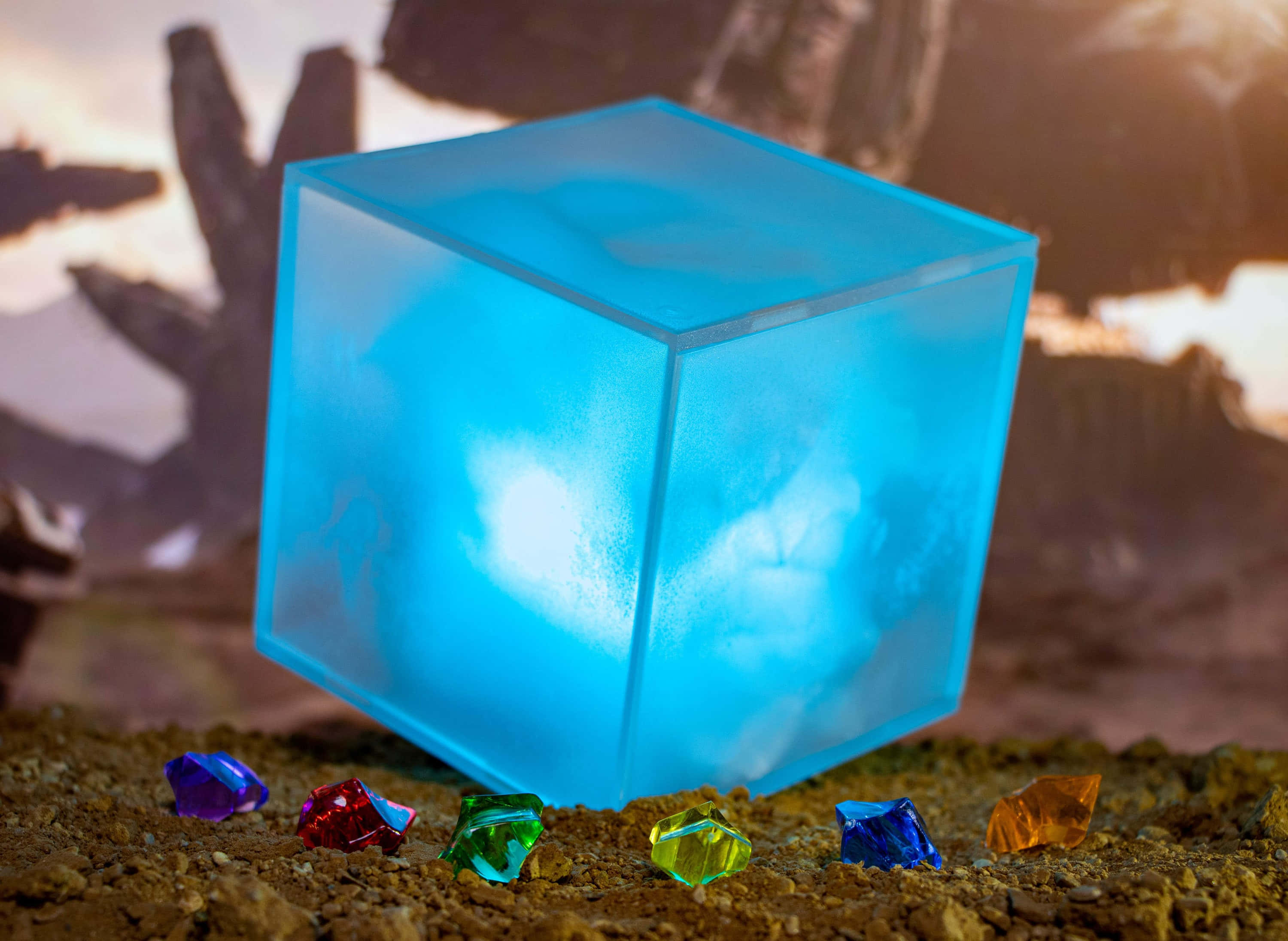 Space Stone 桌布 壁紙 — 一個發光的藍色立方體發出柔和的光線，揭示內部的雲朵，而色彩繽紛的寶石則安放在泥土地上，背景是岩石。