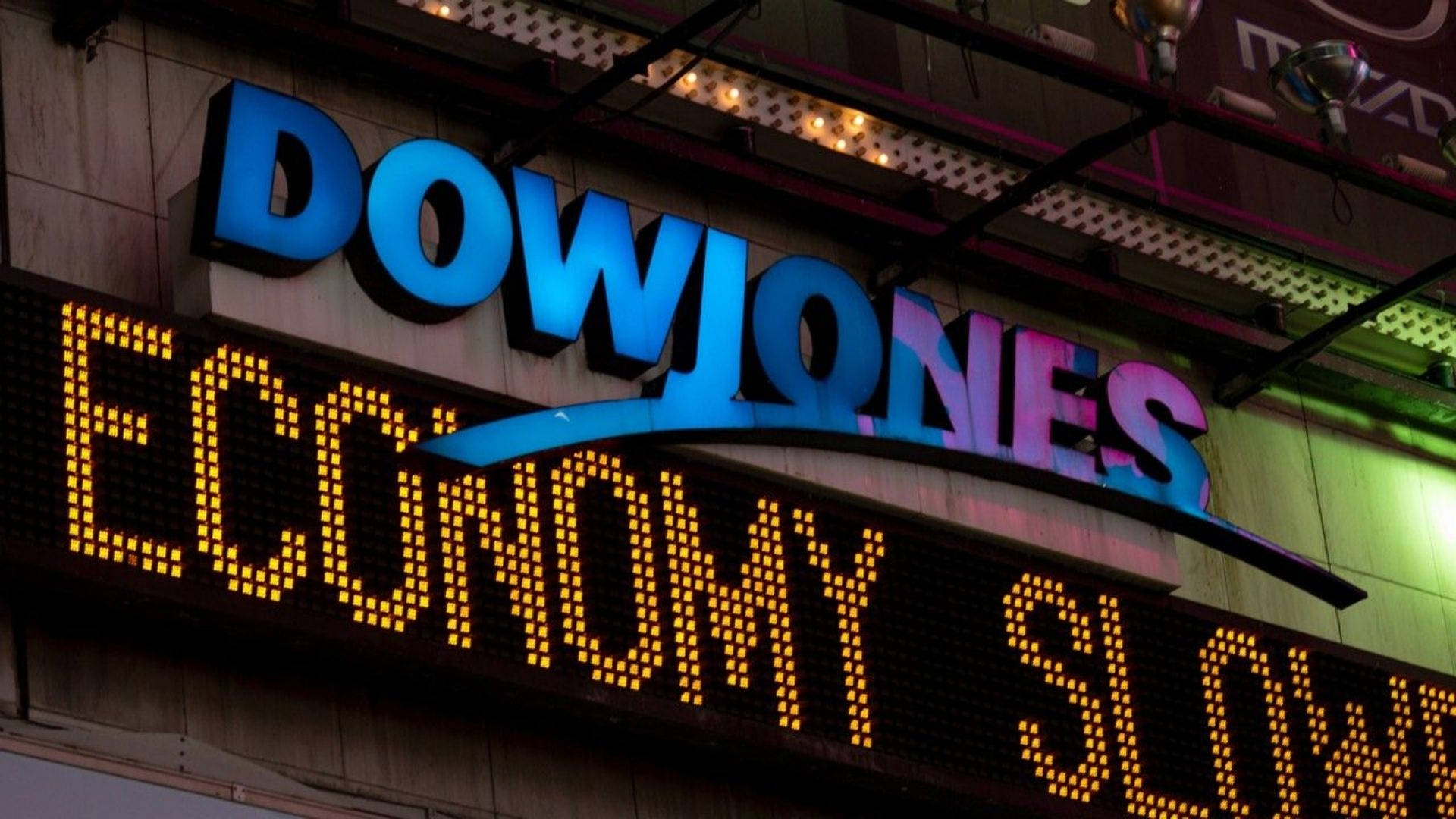 Dow 桌布 壁紙 — 一個發光的藍色道瓊斯標誌，帶有黃色LED跑馬燈顯示economy slow，背景是黑暗的城市建築外牆。