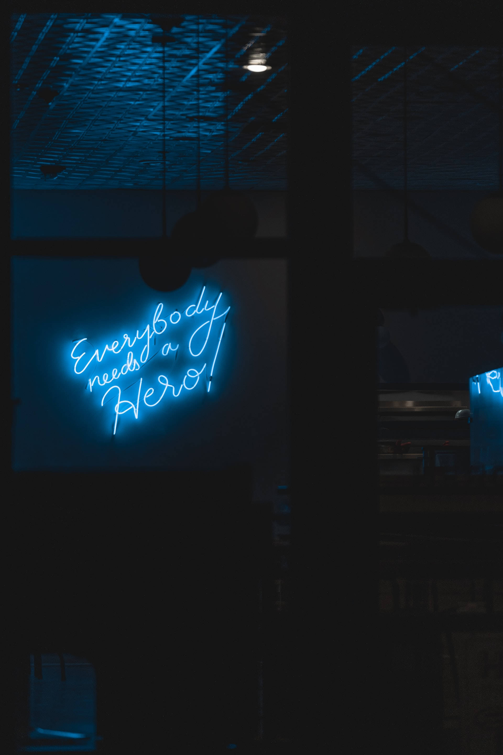 Neon Lights Aesthetic 桌布 壁紙 — 一個發光的藍色霓虹燈牌，上面寫著「Everybody needs a Hero!」，照亮了一個黑暗的室內空間，上方有紋理天花板。